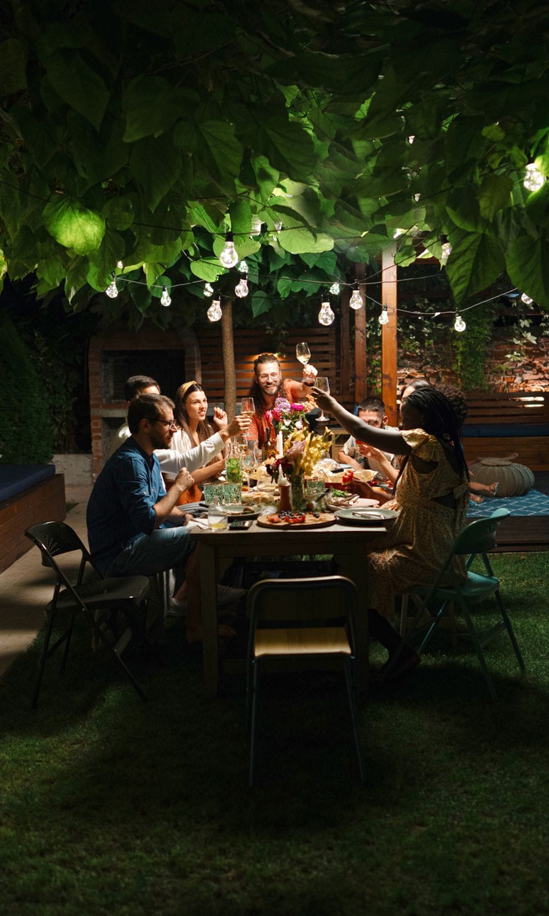 Un grupo de amigos y familiares celebrando en el jardín de una casa en una mesa con guirnalda de luces por la noche.