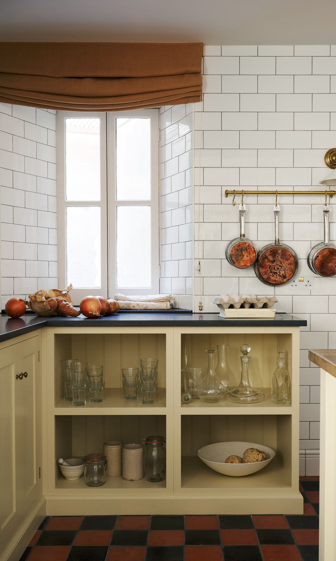 Cocina con pared de azulejos blancos, suelo de damero en marrón y negro, estor marrón, sartenes colgando en una barra, encimera azul, muebles color crema abiertos