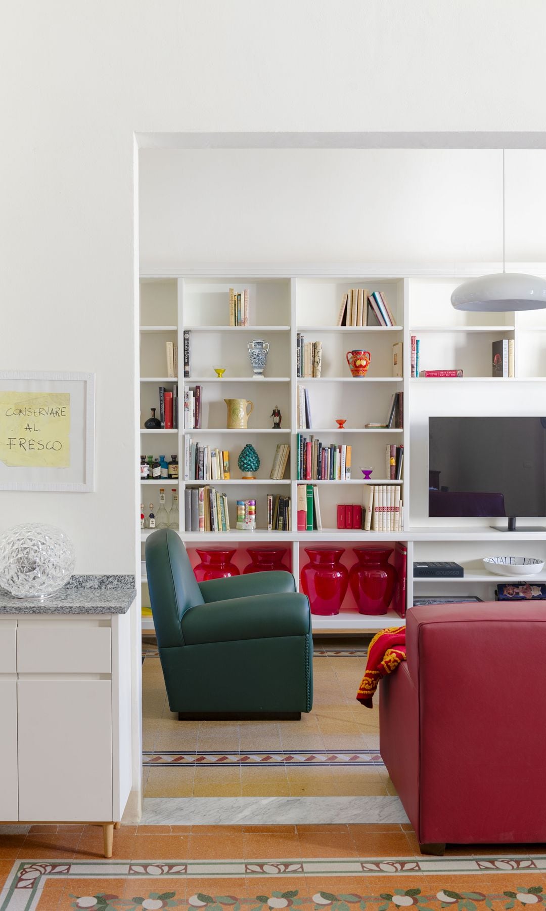 Salón con sofás de cuero rojo, suelos hidráulicos originales, butaca de cuero verde, estantería de madera blanca, con libros y adornos, vasijas de cristal rojo
