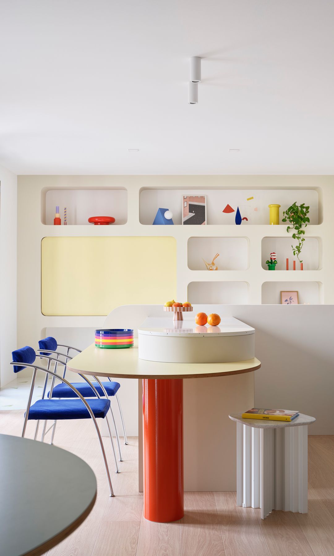 Salón comedor con mesa curva con pie rojo, sobre amarillo, sillas azules, estantería de obra, adornos de colores