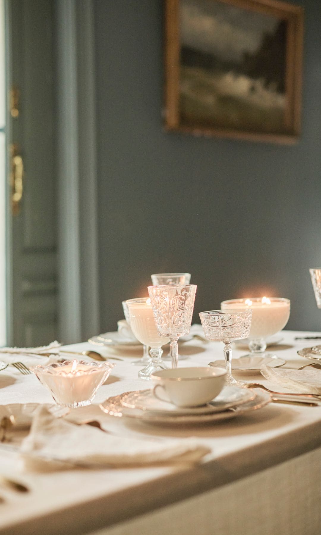Mesa de comedor vestida con mantel, velas encendidas, vajilla blanca con el canto dorado, servilletas de tela, copas labradas