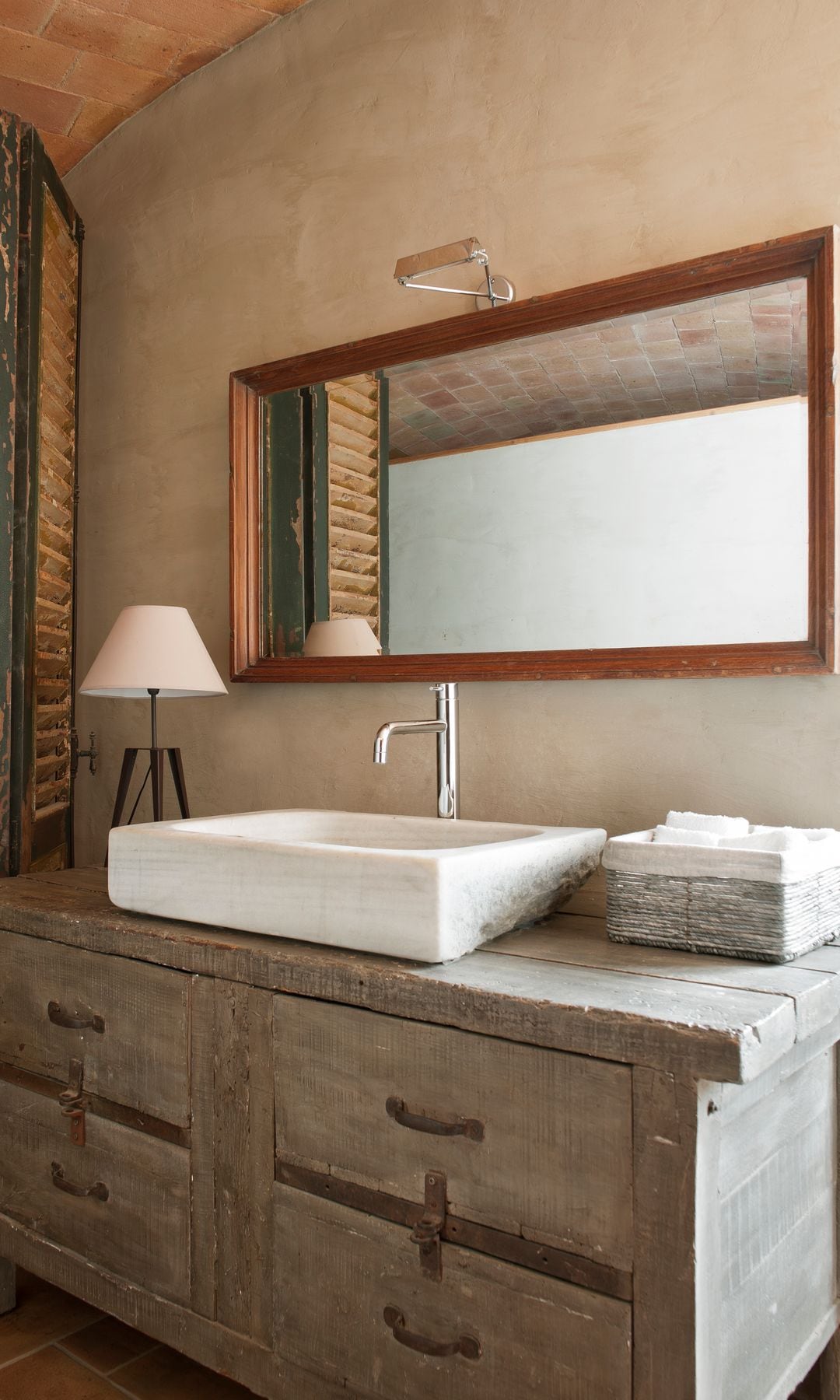 Baño con mueble de madera antiguo restaurado, lavabo sobre encimera, espejo, persianas de madera, lámpara de sobremesa 