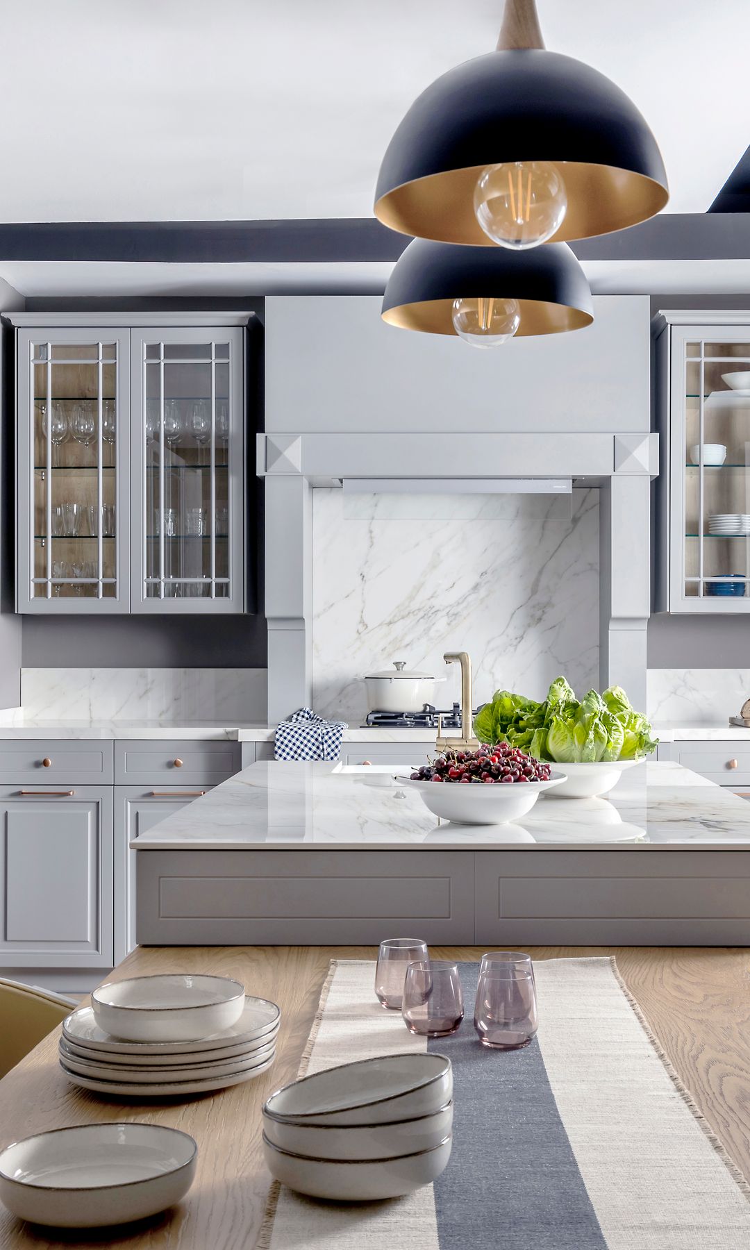 Cocina de estilo inglés con muebles grises, vitrinas superiores, isla con mesa de madera a continuación, lámpara de techo negras