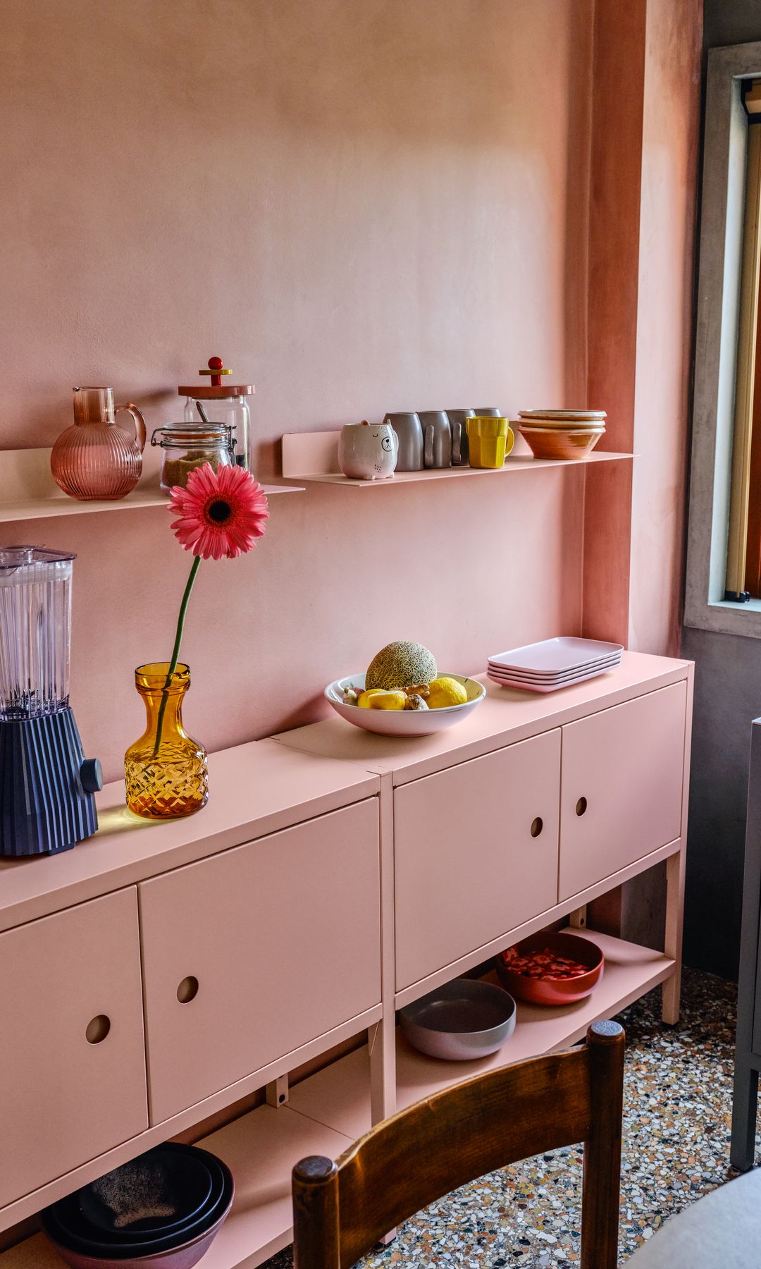 Cocina con paredes y mueble rosa claro, balda de color rosa, jarrón amarillo de cristal con flor roja, paltos y tazas de colores