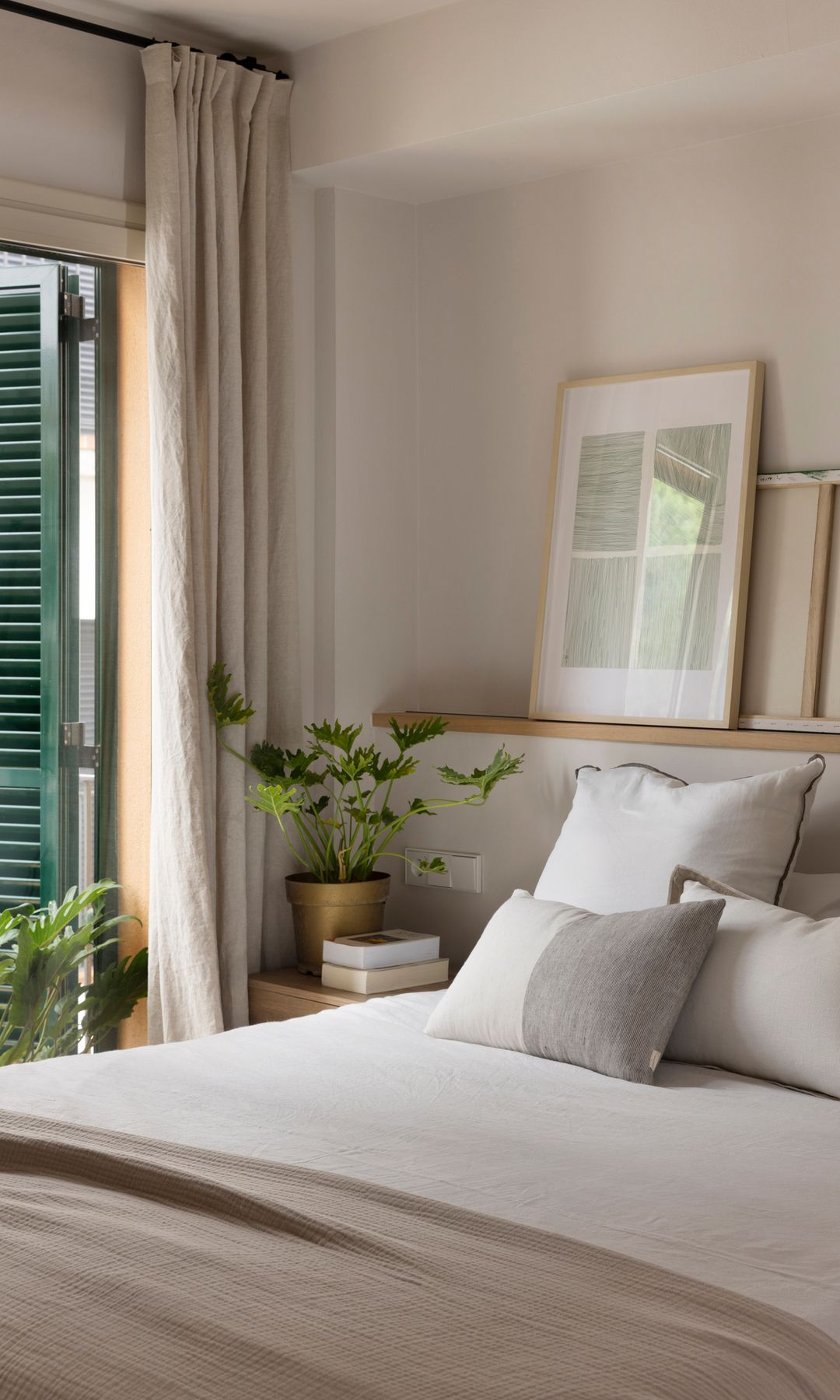 Dormitorio con cabecero de obra con balda de madera, cuadros, ropa de cama beige, plantas, cortinas