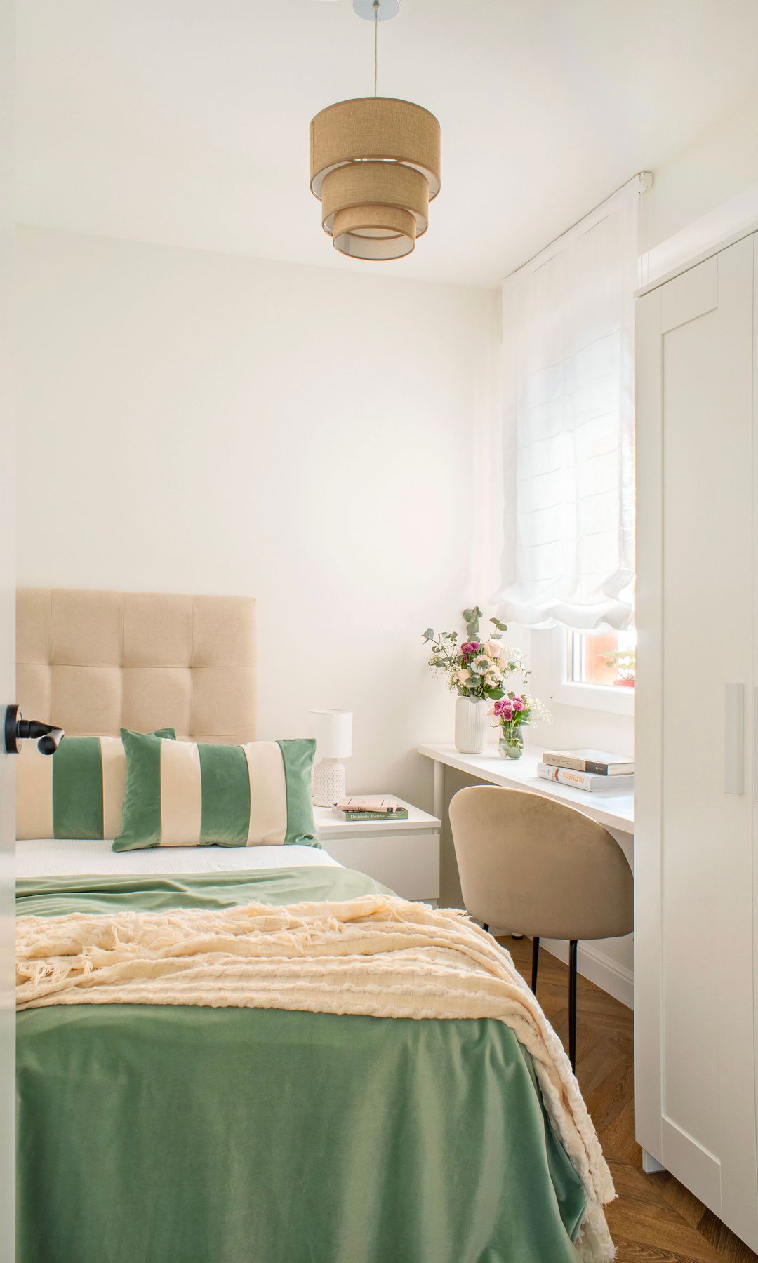 Dormitorio con cabecero tapizado, lámpara de techo, ropa de cama verde, escritorio bajo la ventana con silla beige, estor blanco