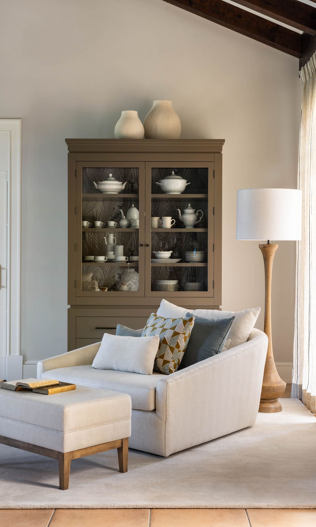 Rincón de lectura en el salón con vitrina de madera con vajilla blanca, lámpara de pie, sillón beige y reposapiés a juego