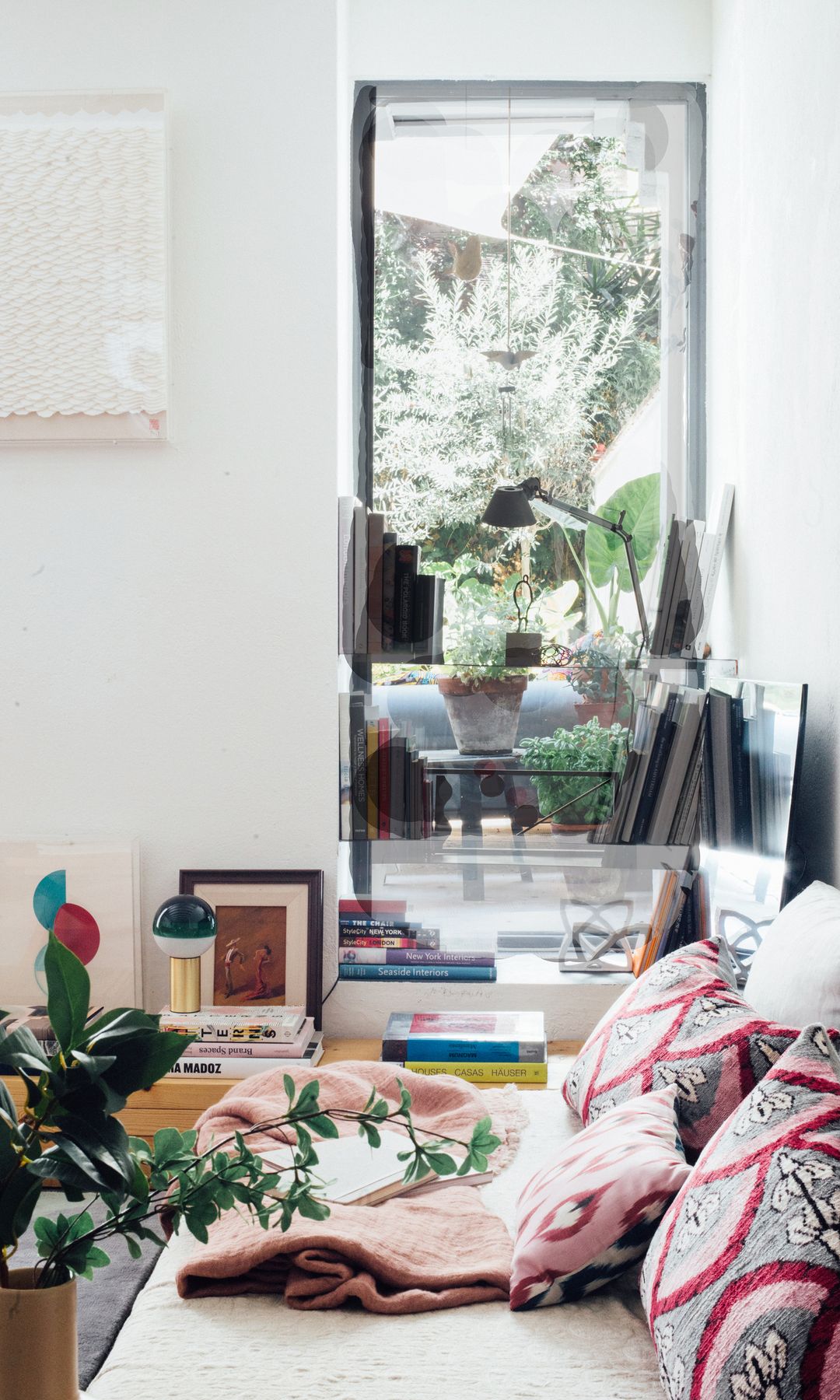 Salón con sofá gris con cojines rosas, ventanal con salida al patio, plantas, libros 