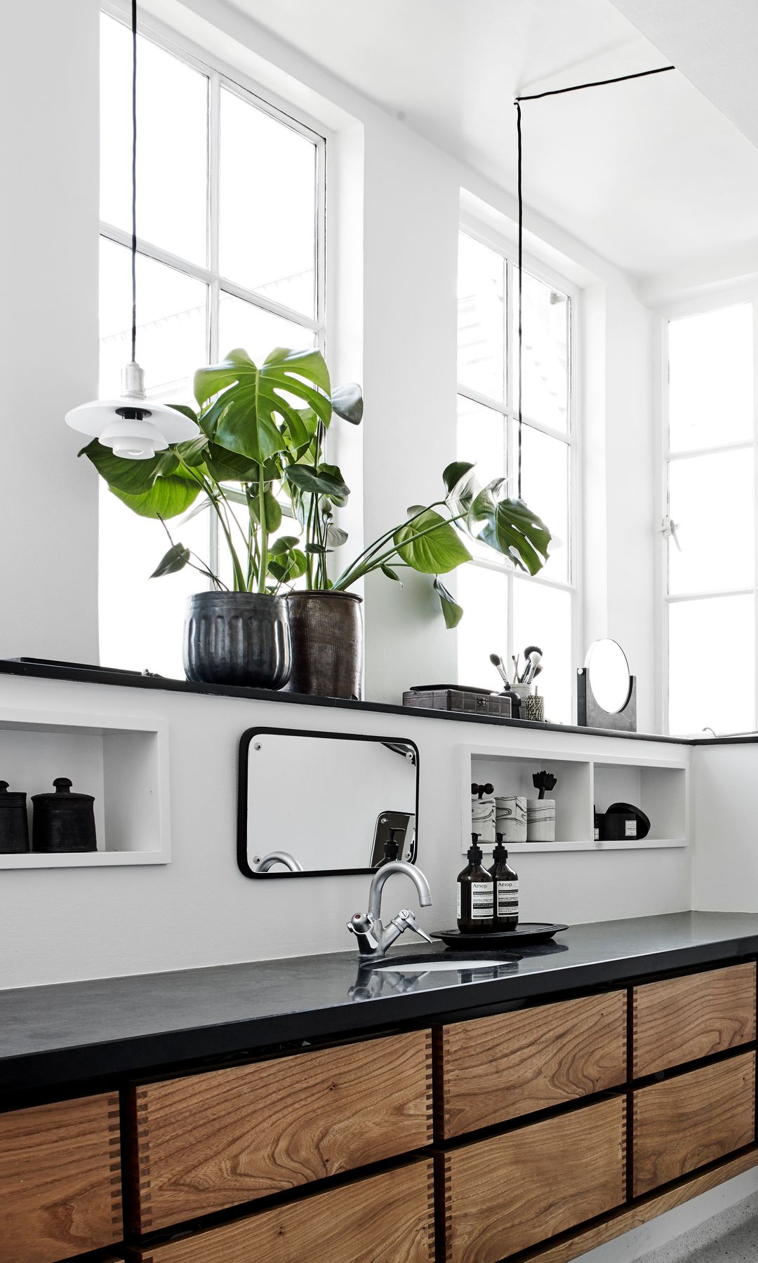 Baño con espejo redondo, hornacinas, mueble de baño de madera, espejo en el suelo, plantas en macetas negras, alfombra