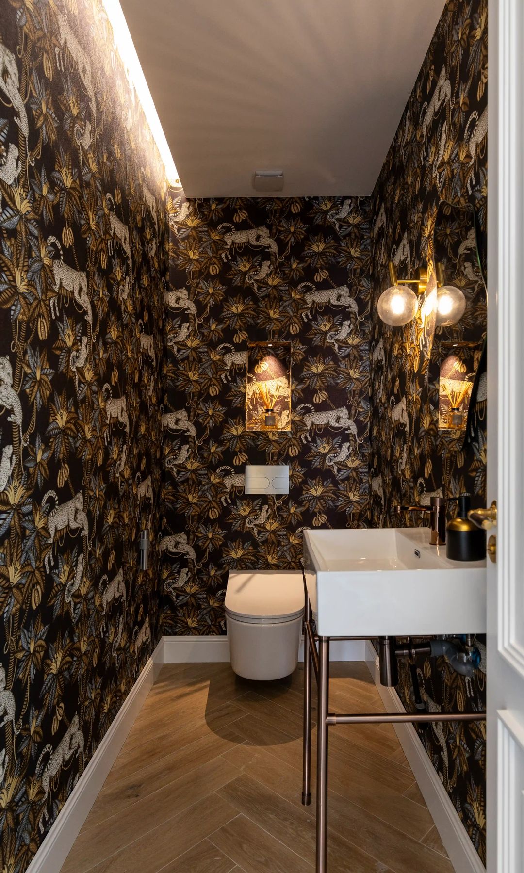 Aseo de cortesía con papel pintado negro, techo retroiluminado, lavabo con patas metálicas, suelo de madera y lámparas doradas