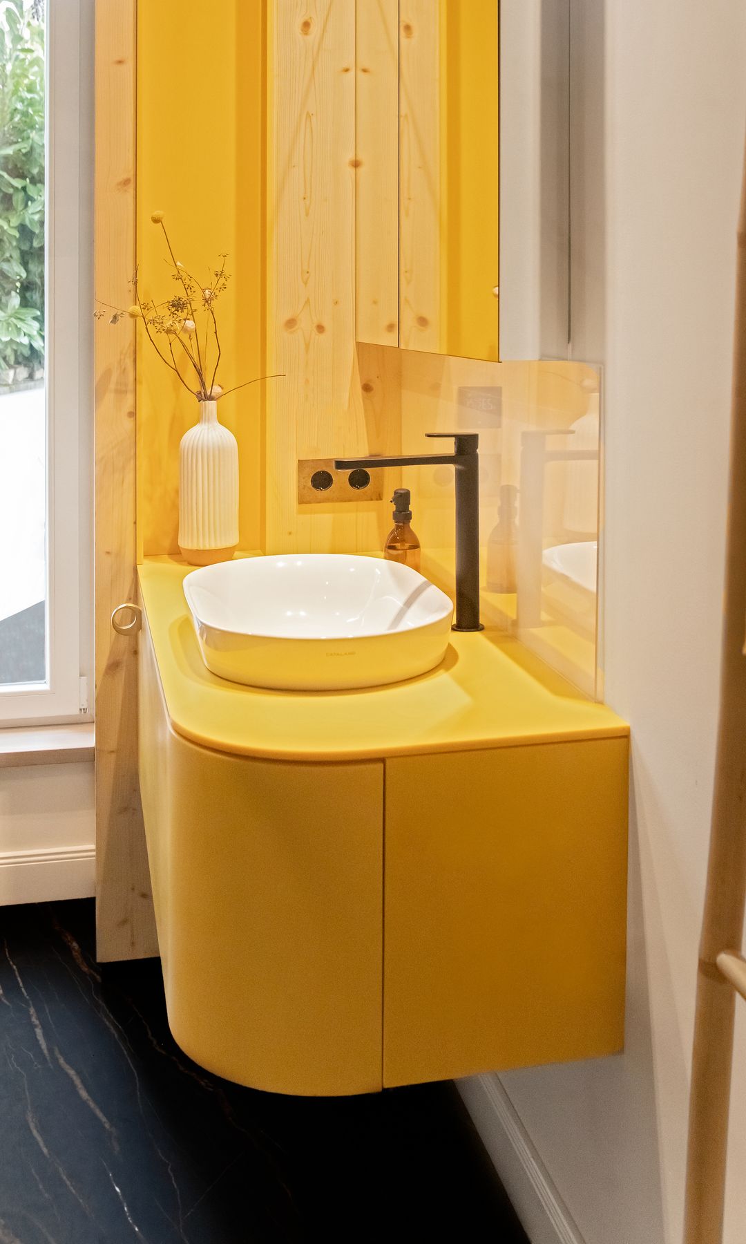 Baño con mueble bajaolavabo amarillo suspendido, mueble amarillo, lavabo blanco, suelo negro y grifería negra