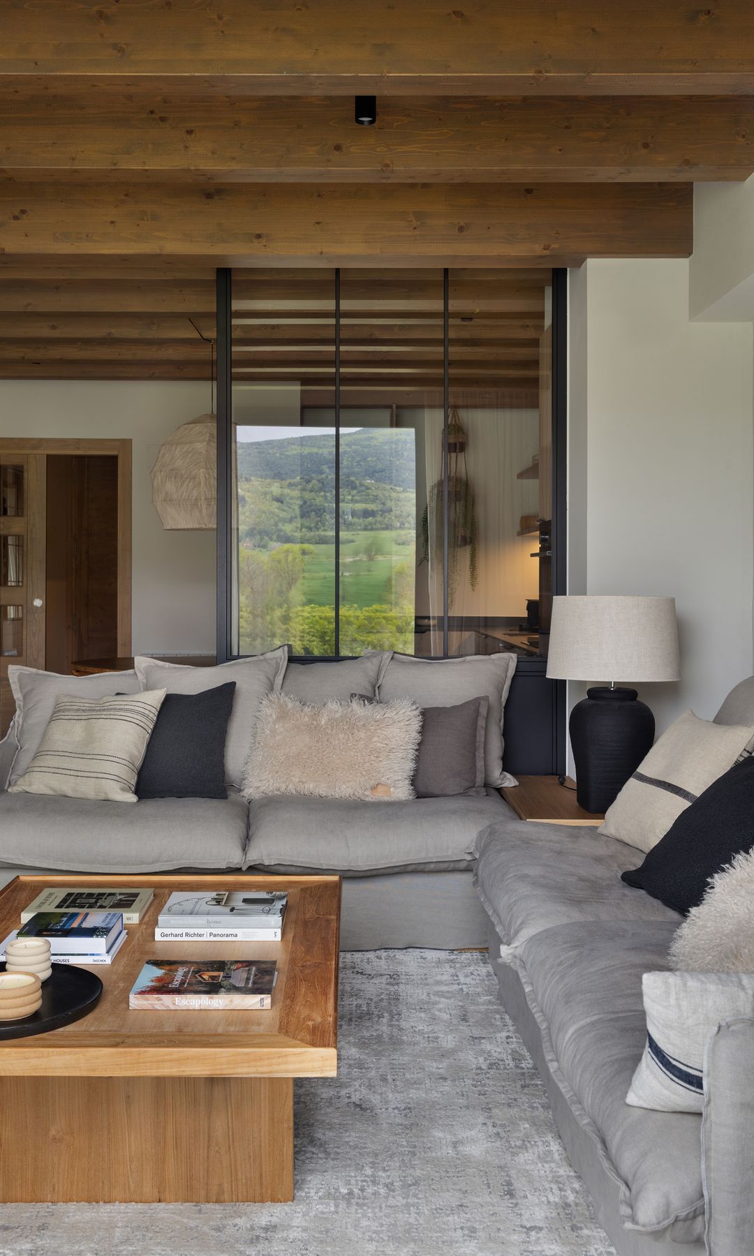 Salón con alfombra de pelo largo gris, sofás grises, cojines blancos, grises y negros, mesa de centro de madera, techo con vigas de madera