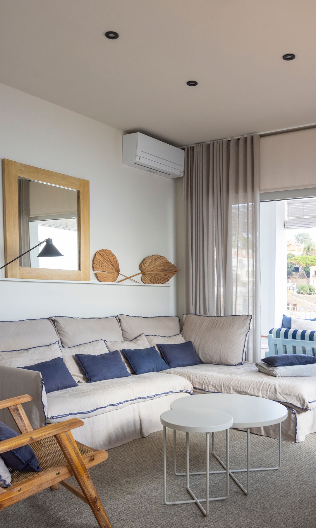 Salón con gran ventanal, sofá blanco de lino, cojines azules, espejo con el marco de madera, mesa de centro blanca, alfombra, cortinas beiges