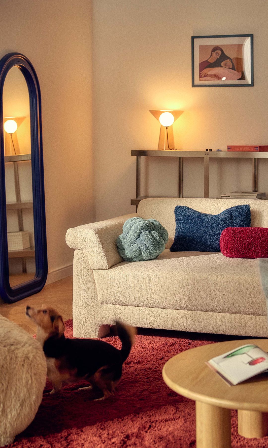 Salón con sofá beige, cojines de colores, alfombra roja, perro en la alfombra, lámpara de sobremesa encendida, espejo de pie