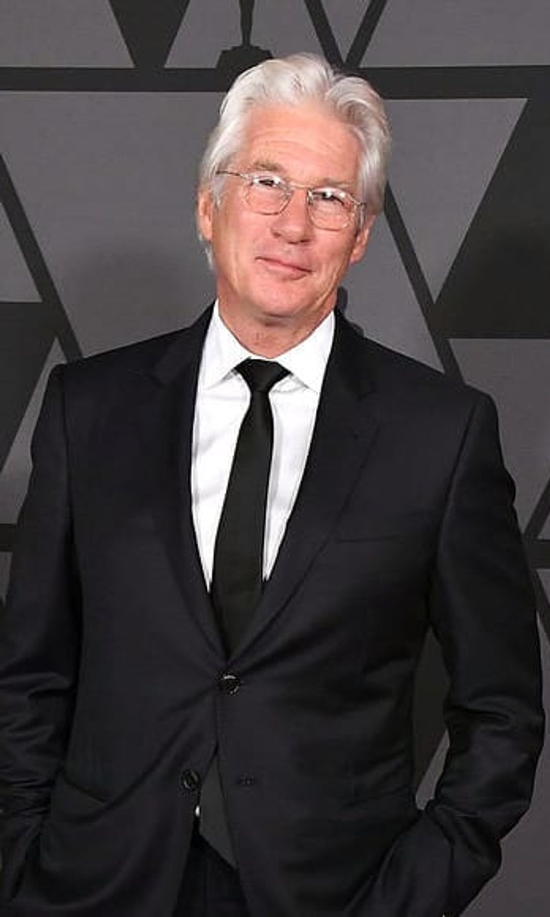 Richard Gere, con cabello blanco y traje negro, posa en una alfombra roja.