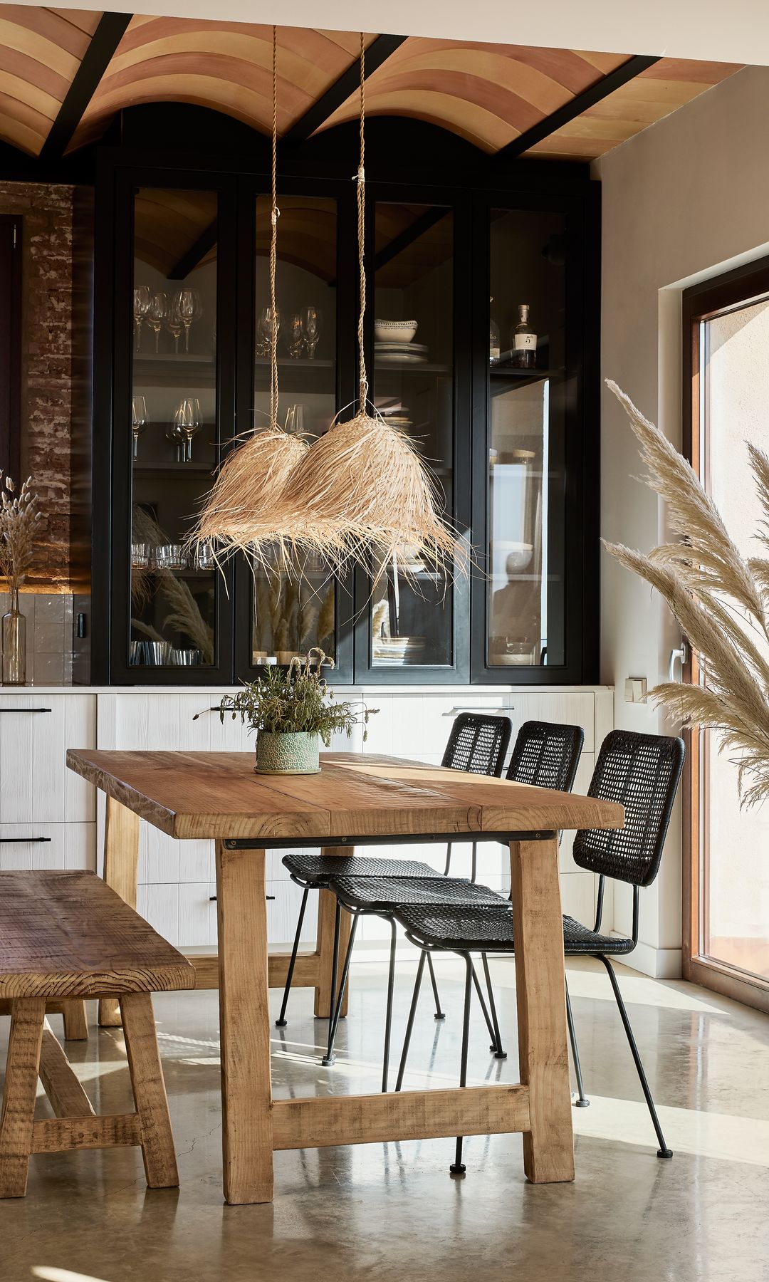 Cocina con office, mesa de madera, banco de madera, sillas negras, bóvedas techo, vitrina negra sobre la encimera, lámparas de techo de fibras vegetales 