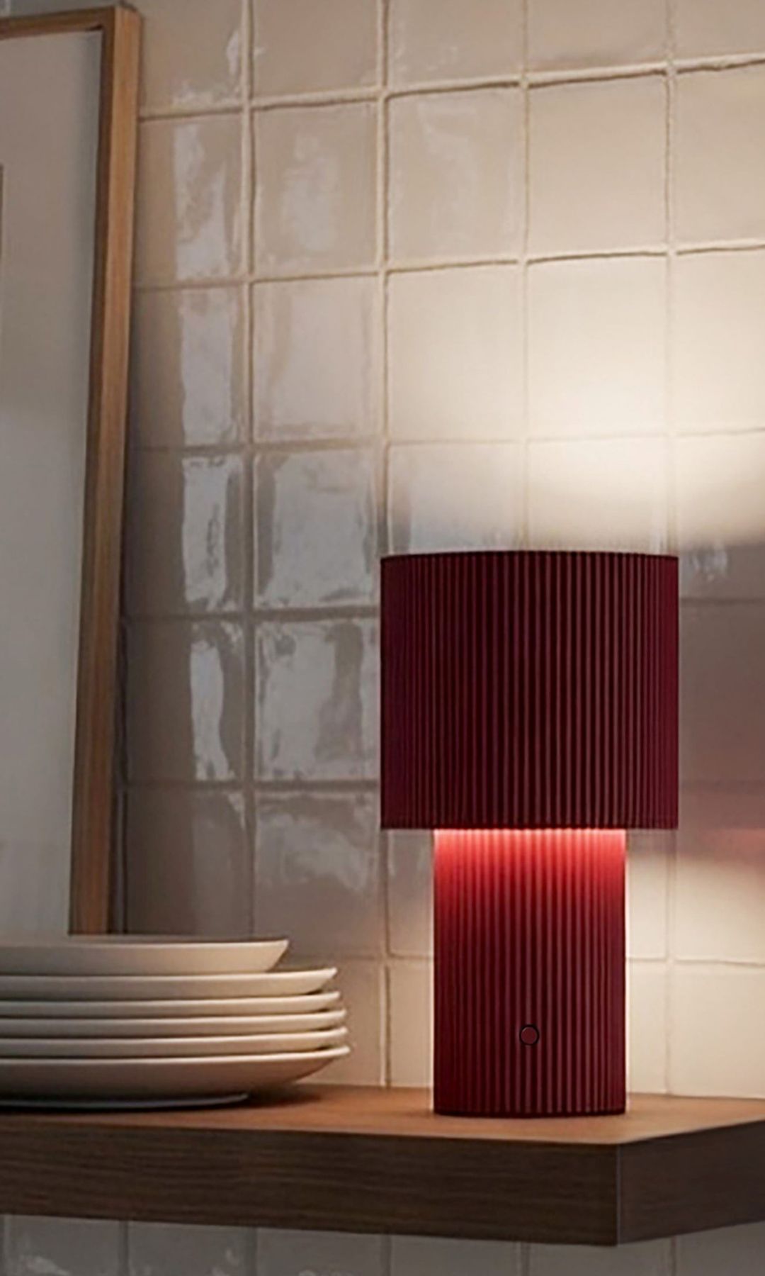 Pared de azulejos con balda de madera, lámpara de sobremesa roja, platos blancos y lámina apoyada