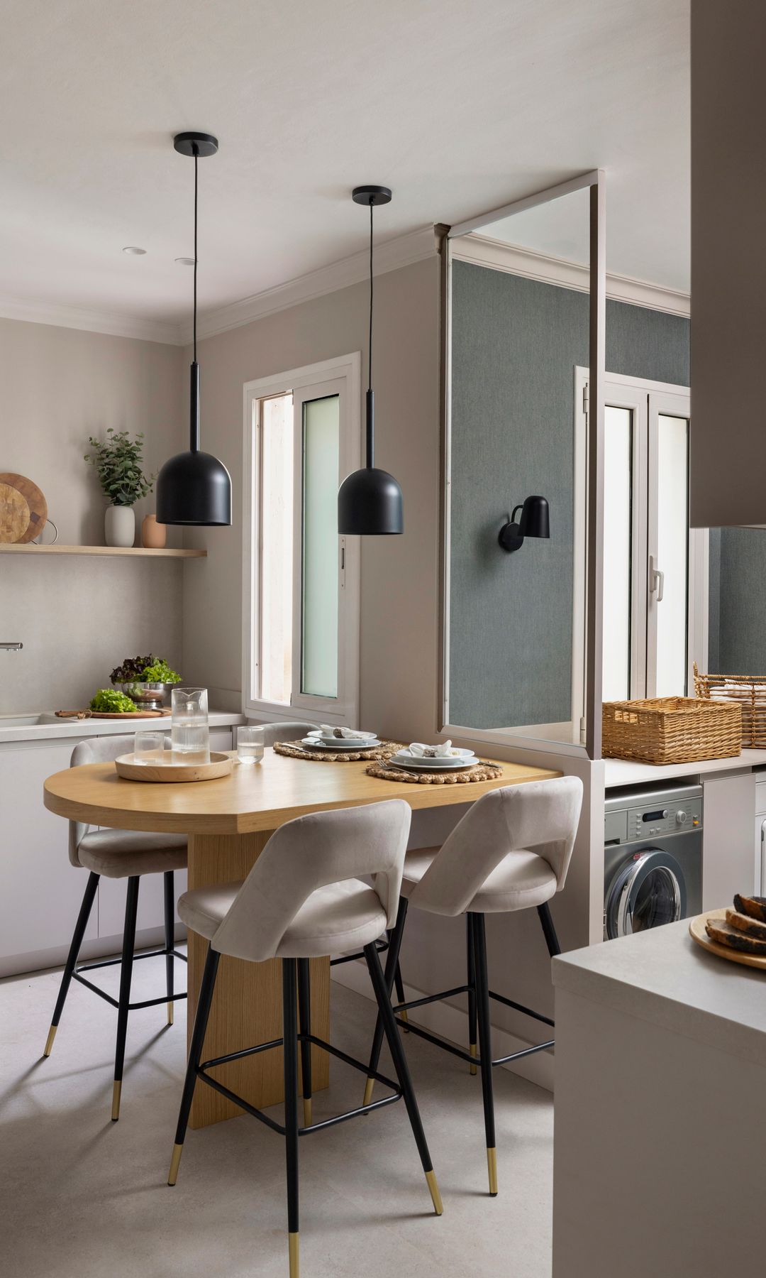 Cocina separada del tendedero mediante tabique de cristal con mesa de offices de madera y taburetes altos tapizados, lámparas de techo negras