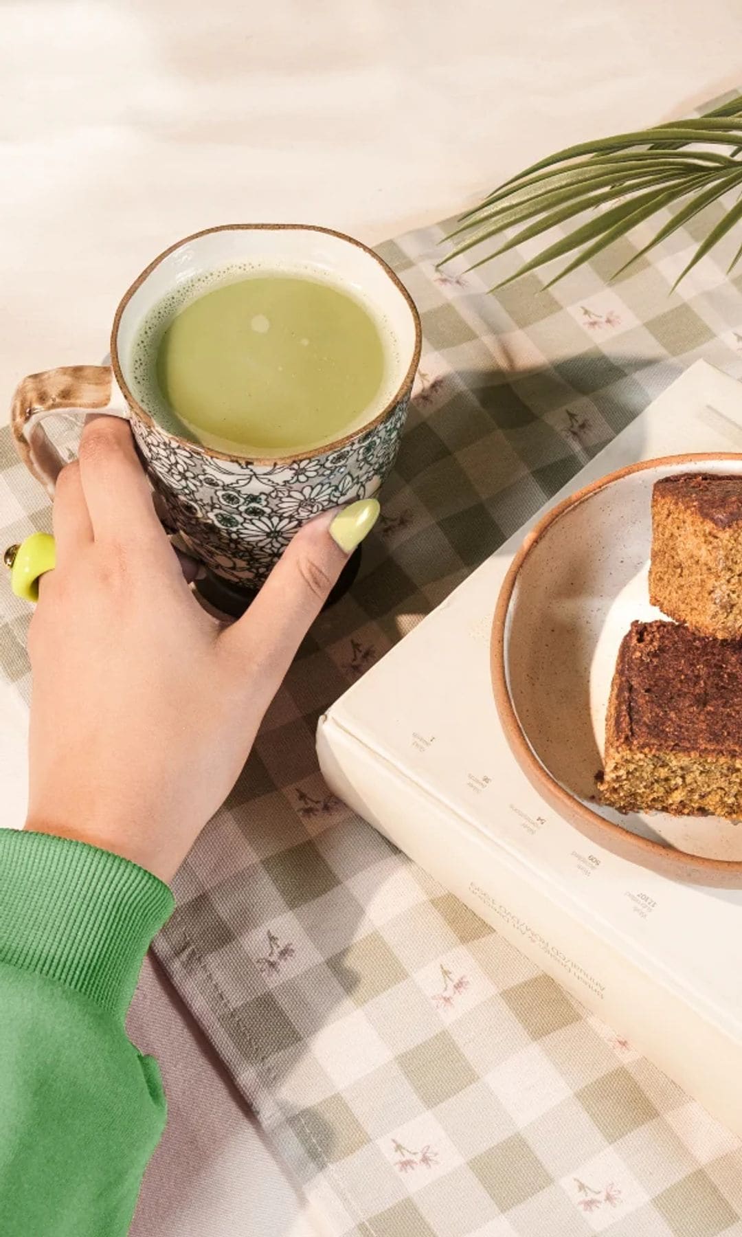 Té matcha con bizcocho en una mesa, sujetando la taza una mujer con jersey verde y uñas verde lima.