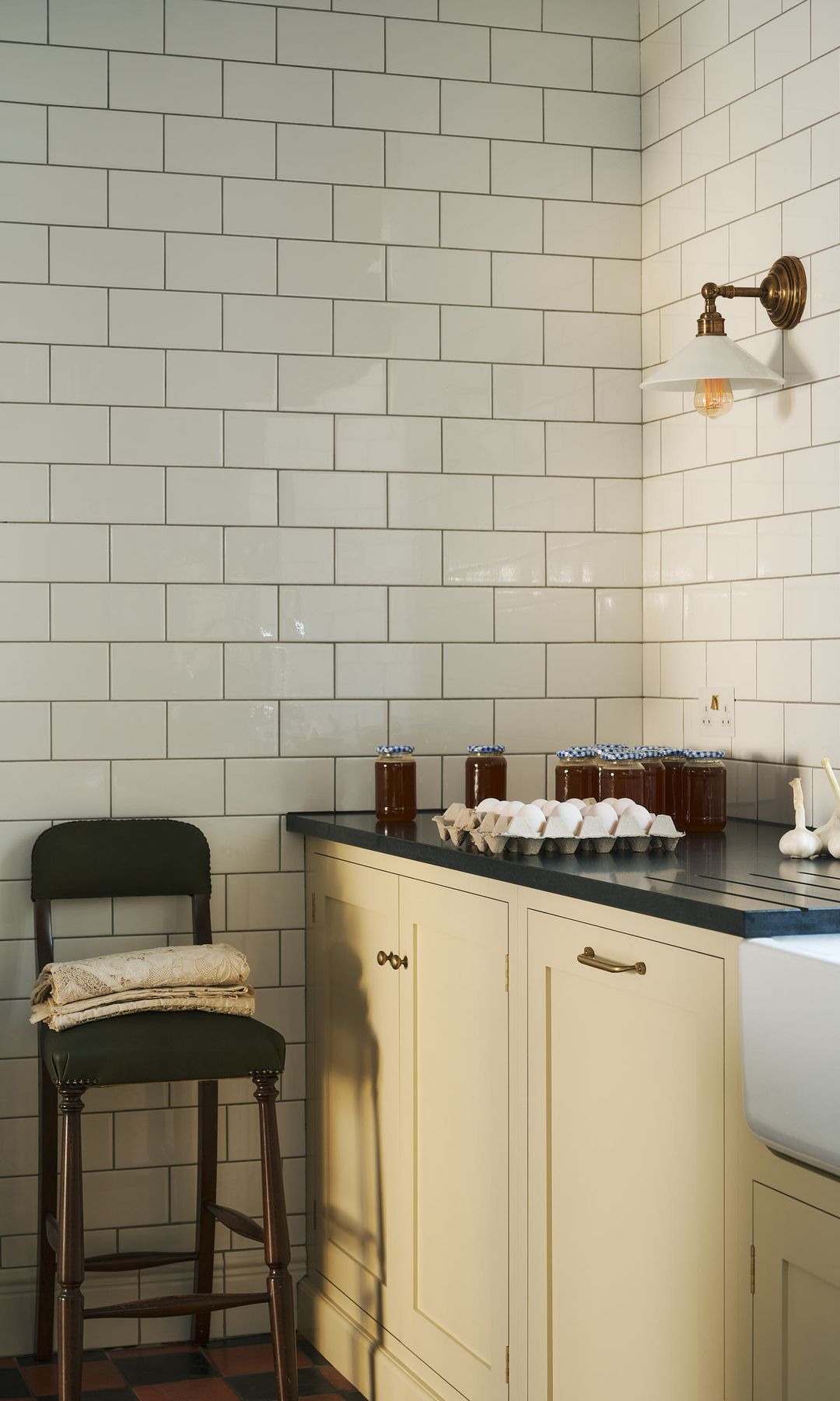 Cocina con muebles de color crema, azulejos blancos brillantes, tipo metro, taburete de madera alto, encimera negra, aplique dorado en la pared, cartón de huevos, ajos y botes de mermelada