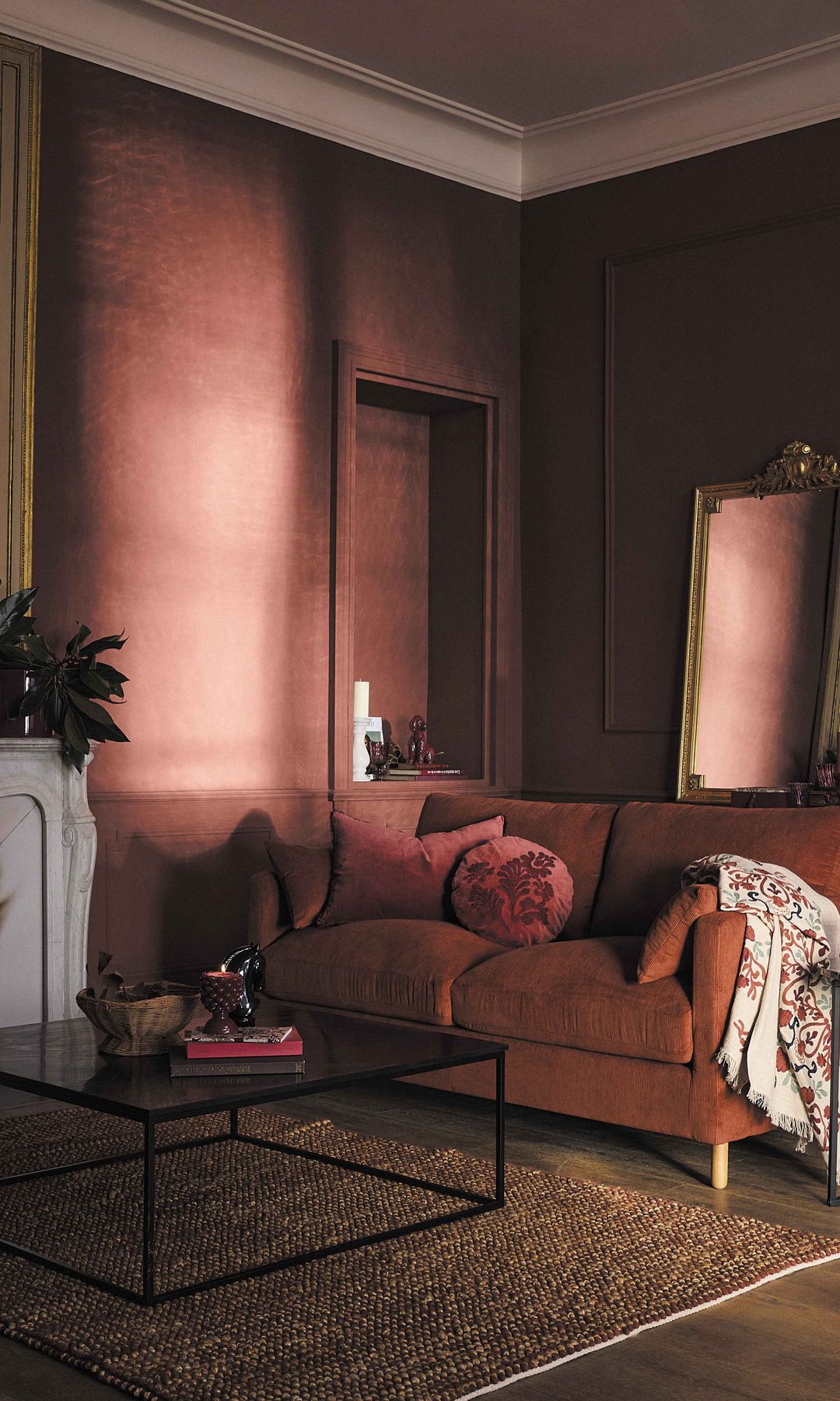 Salón con chimenea, paredes en color marrón, sofá de terciopelo de color chocolate, alfombra, mesa de centro negra, espejo con el marco dorado apoyado en un aparador, cojines, plaid sobre el brazo del sofá