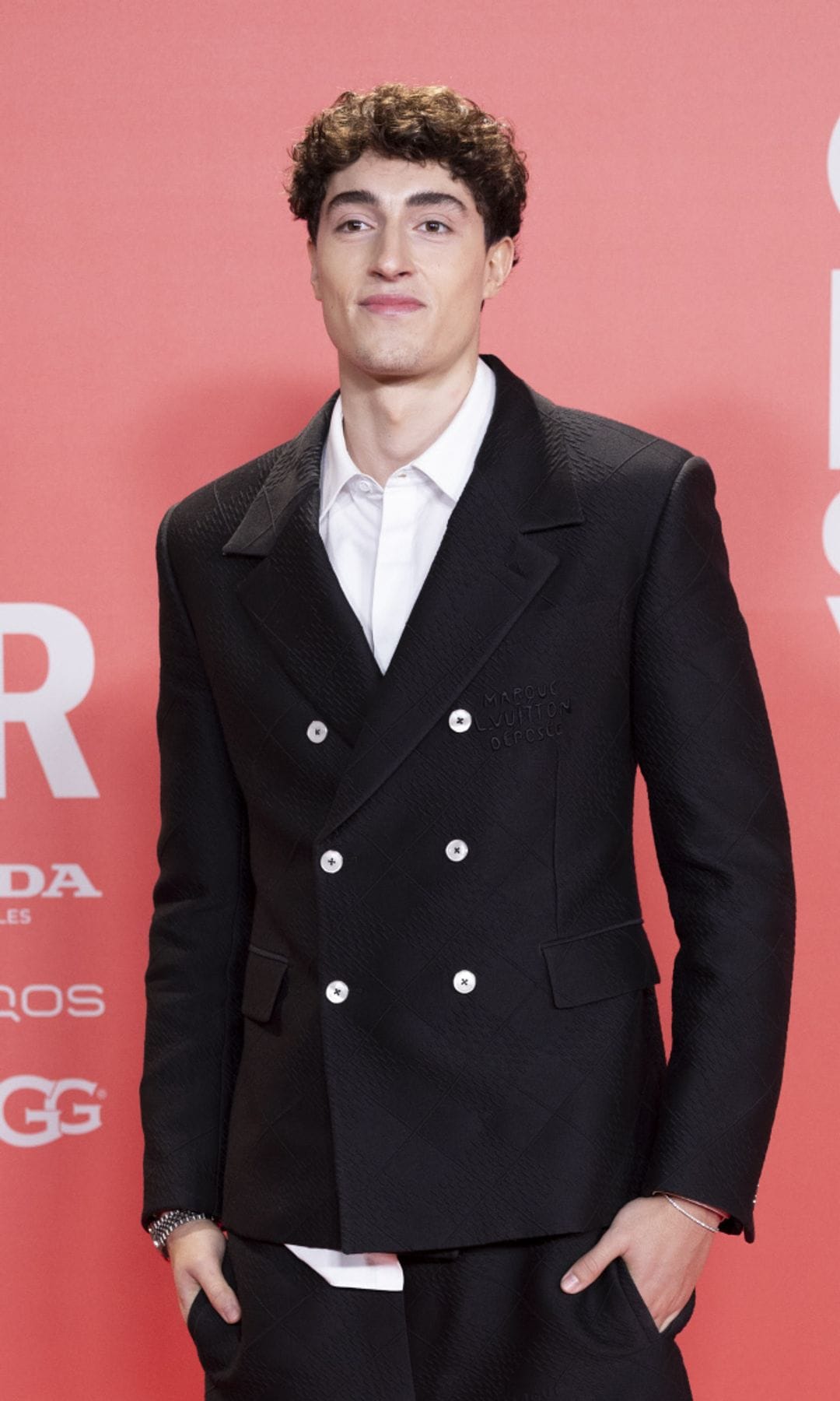 Plex en traje negro con manos en los bolsillos en una alfombra roja.