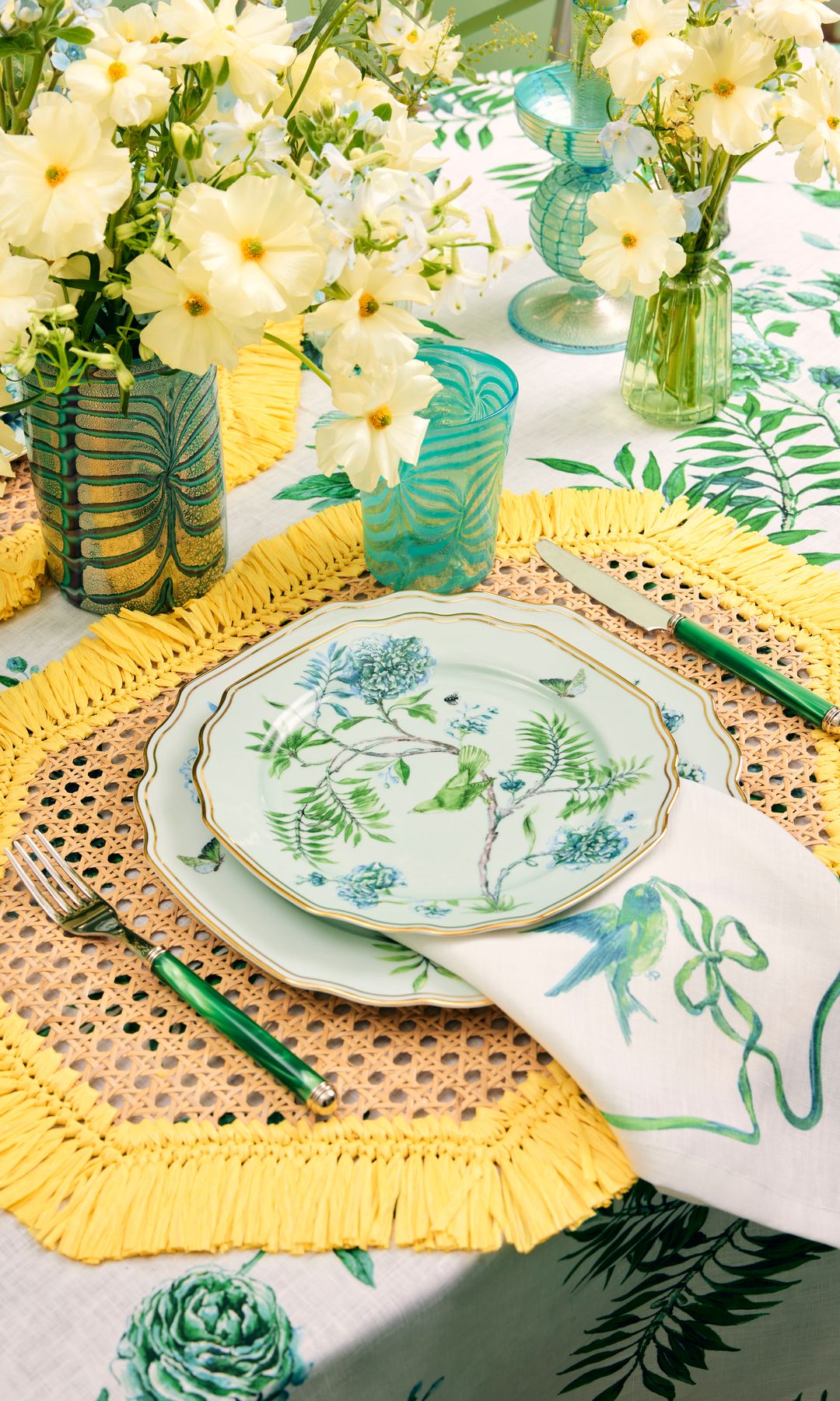 Mesa con individual de rejilla con flecos amarillos, platos con flores verdes, cubiertos y vasos verdes y centro de mesa de flores amarillas