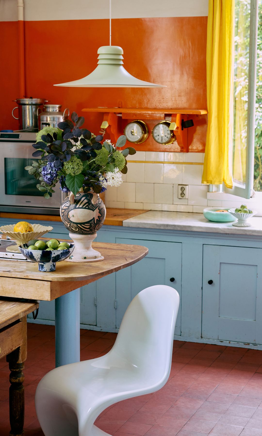 Cocina con paredes naranjas, cortinas amarillas, muebles de cocina azule claro, suelo de barro, mesa de madera, silla blanca, jarrón con flores, lámpara de techo