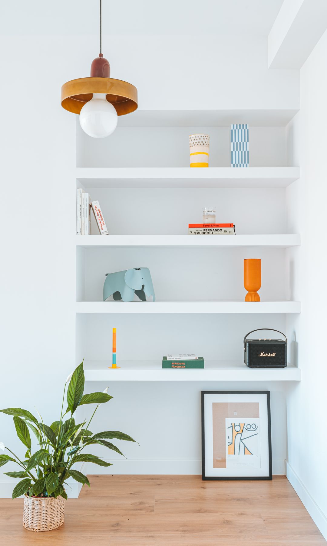 Estantería de obra con baldas con libros y adornos, planta en el suelo, suelo de madera, cuadro en el suelo, lámpara de techo