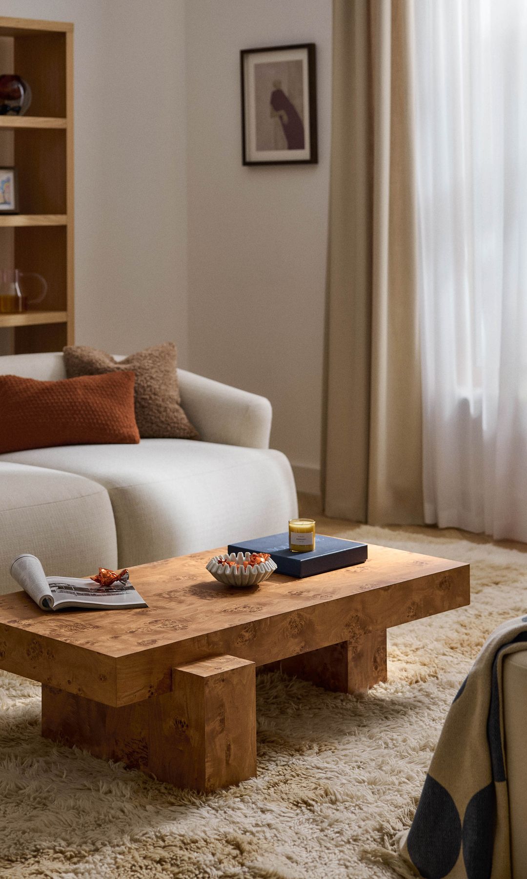 Salón con cortinas y visillos blancos, alfombra de pelo largo, mesa de centro de madera, sofá blanco, cojines en tonos marrones, estantería de madera 