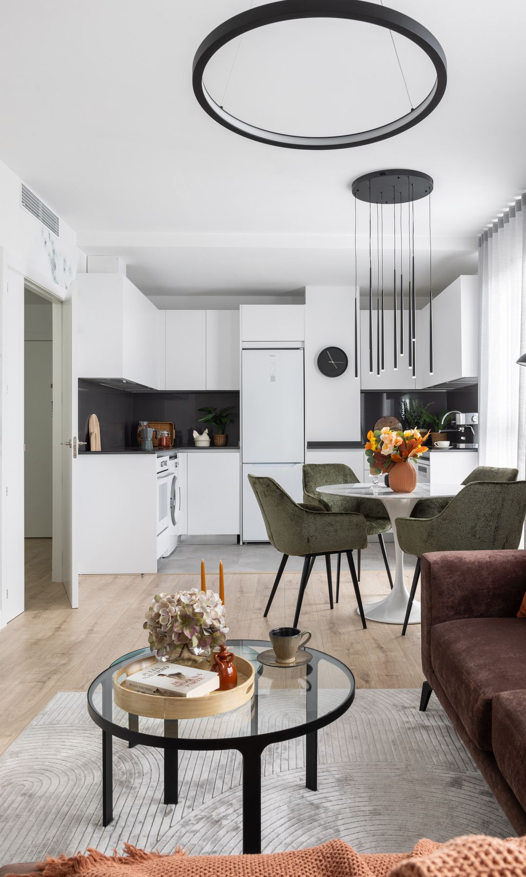 Espacio único con salón, comedor y cocina, suelo de madera, lámparas de techo, muebles de cocina blancos, sofá, mesa de centro de cristal y mesa de comedor redonda
