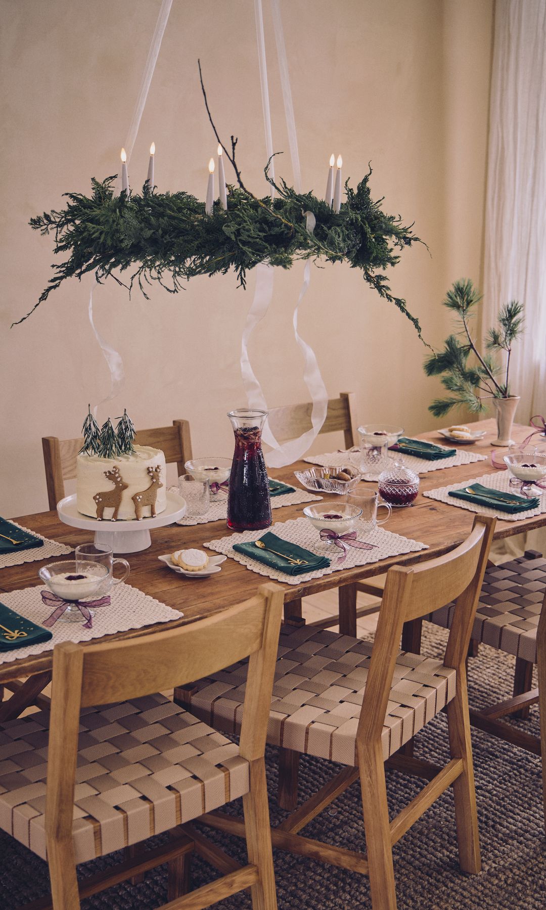 Mesa de Navidad en madera con lámpara de techo decorada con ramas de abeto, sillas de enea, cesto de flores, manteles individuales, servilletas verdes 