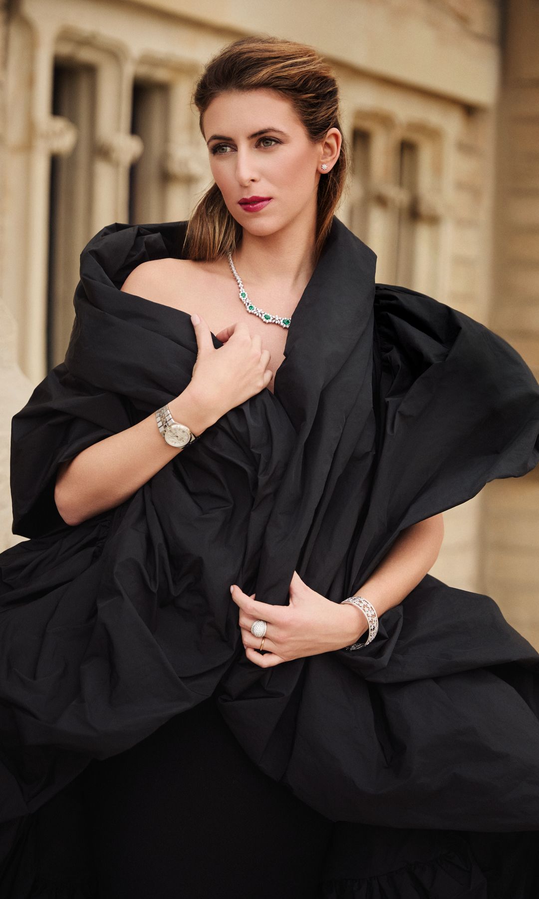 Mujer con cabello corto, vestido negro, joyas y reloj posando en un entorno urbano.