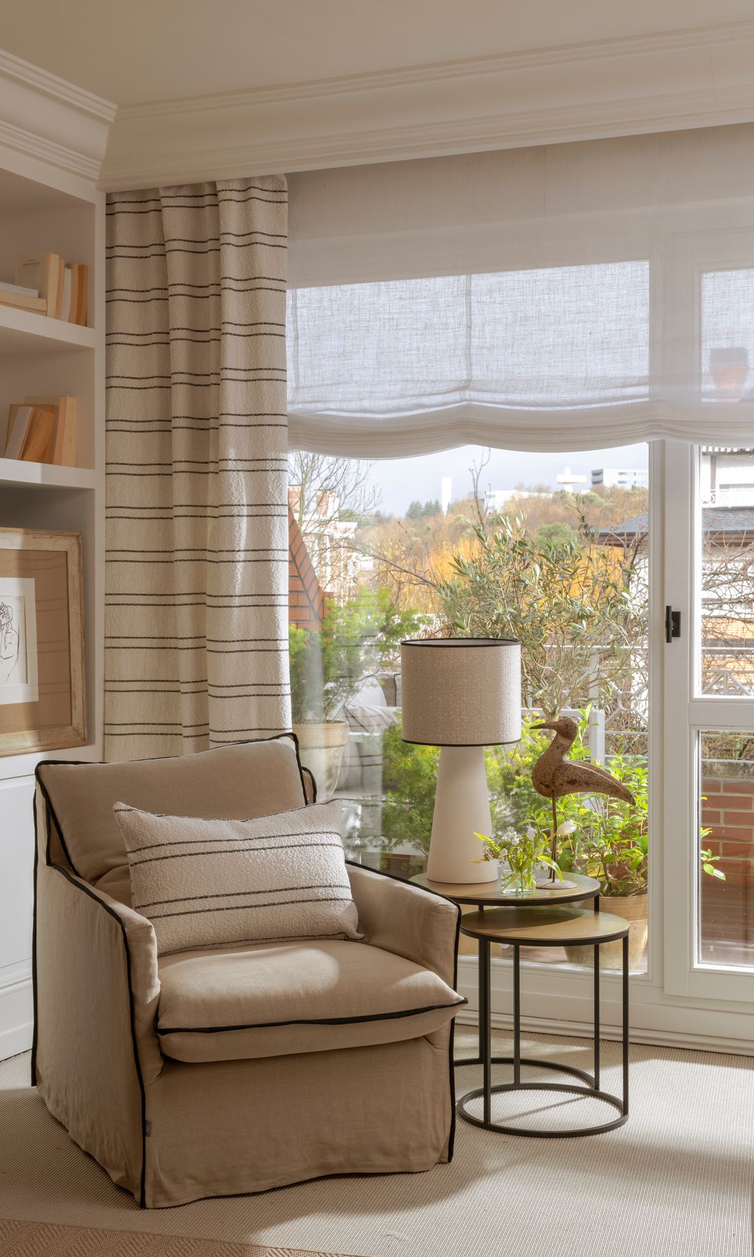 Rincón casa con sillón tipo orejero, mesa auxilia, lámpara de sobremesa, estores blancos, cortinas de rayas, alfombra, salida terraza