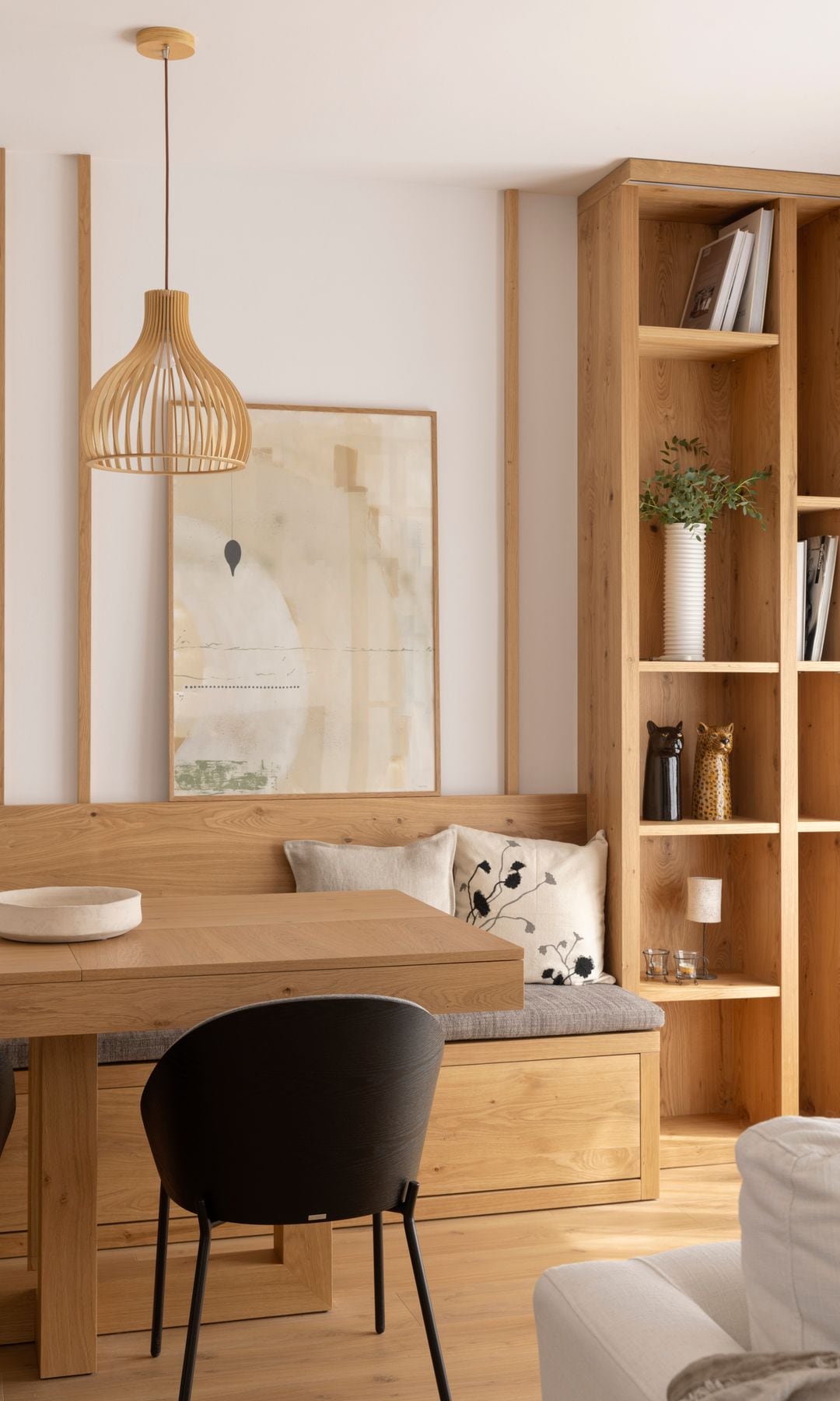 Comedor con banco de madera, librería de madera y mesa a juego, sillas negras, cuadro y lámpara de techo