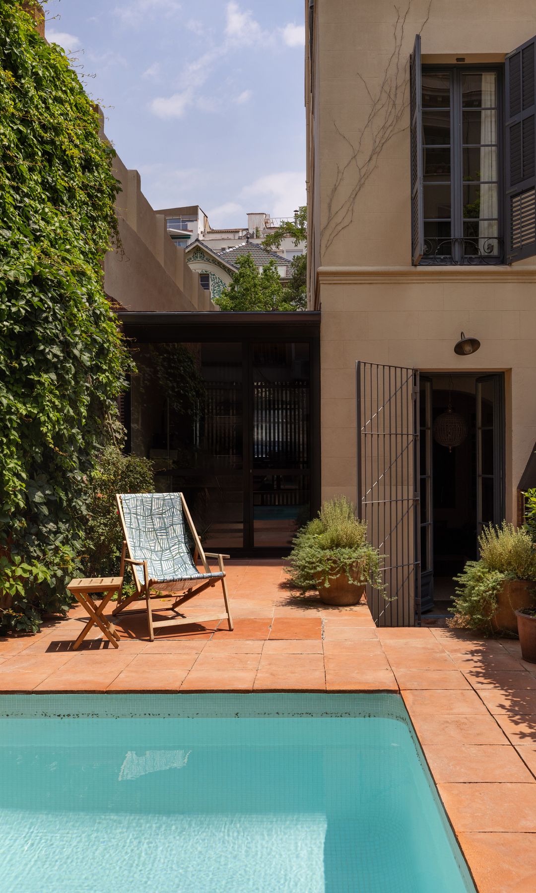 Casa con piscina con suelo de baldosas de barro, hamaca y taburete de madera, macetas con plantas 