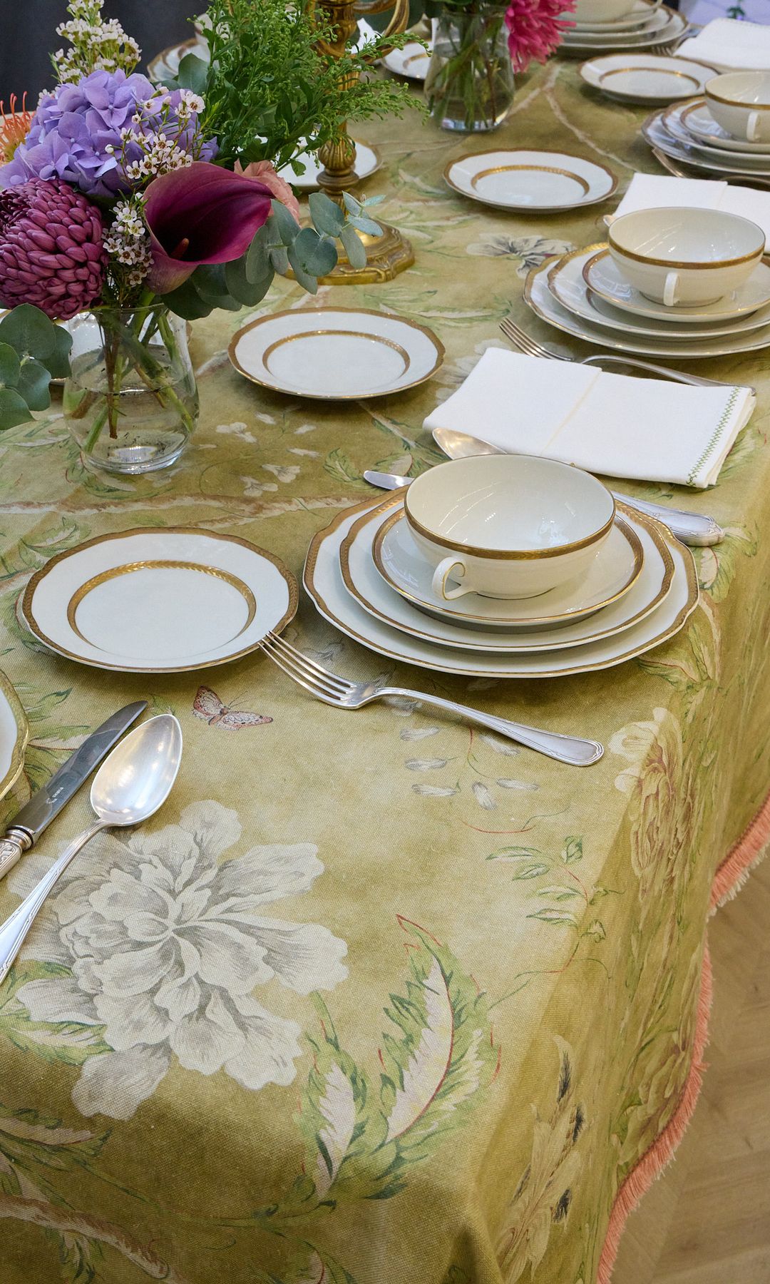 Mesa con mantel verde de flores, vajilla blanca con el filo dorado, cubiertos de plata, jarrón con flores