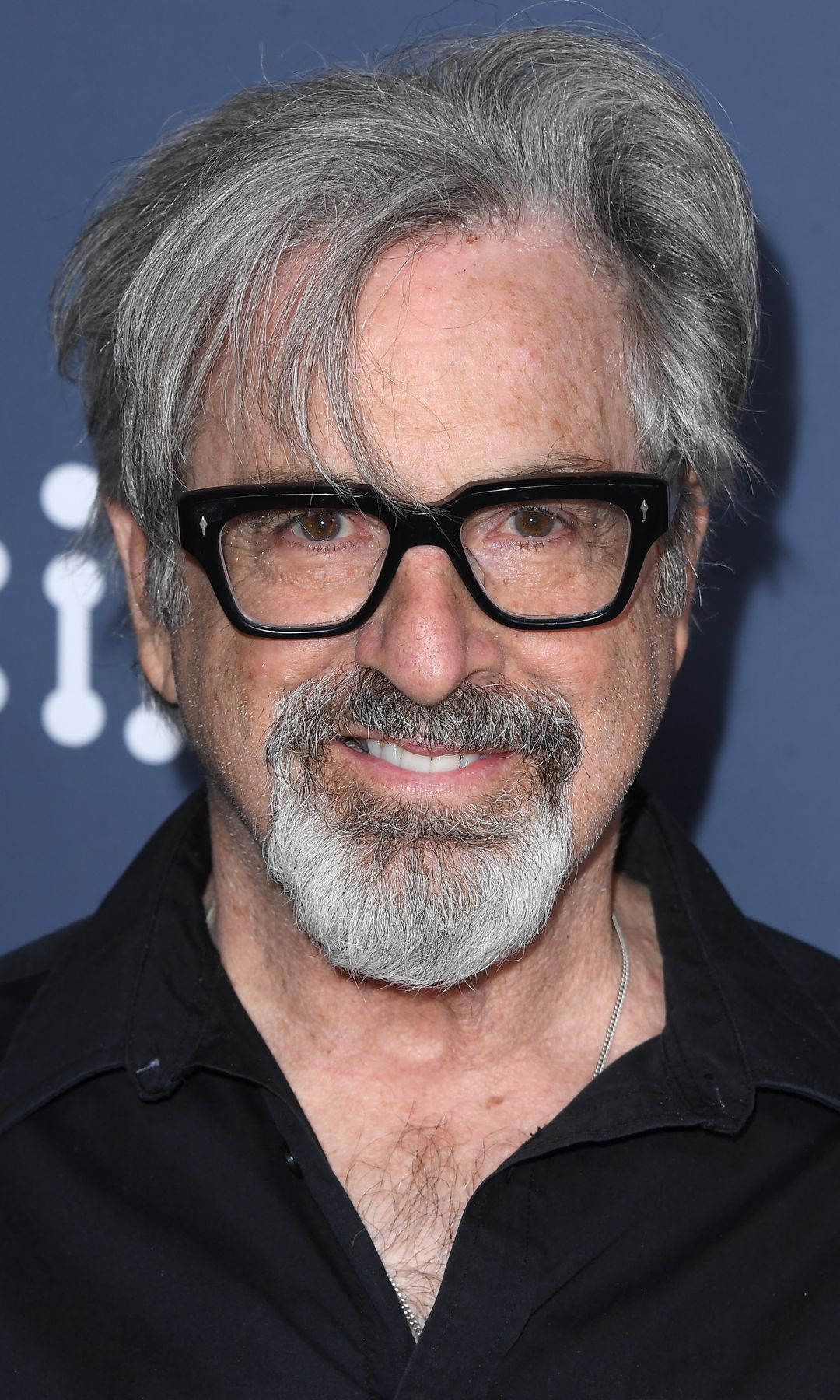 Robert Carradine sonríe, usa gafas negras y camisa negra