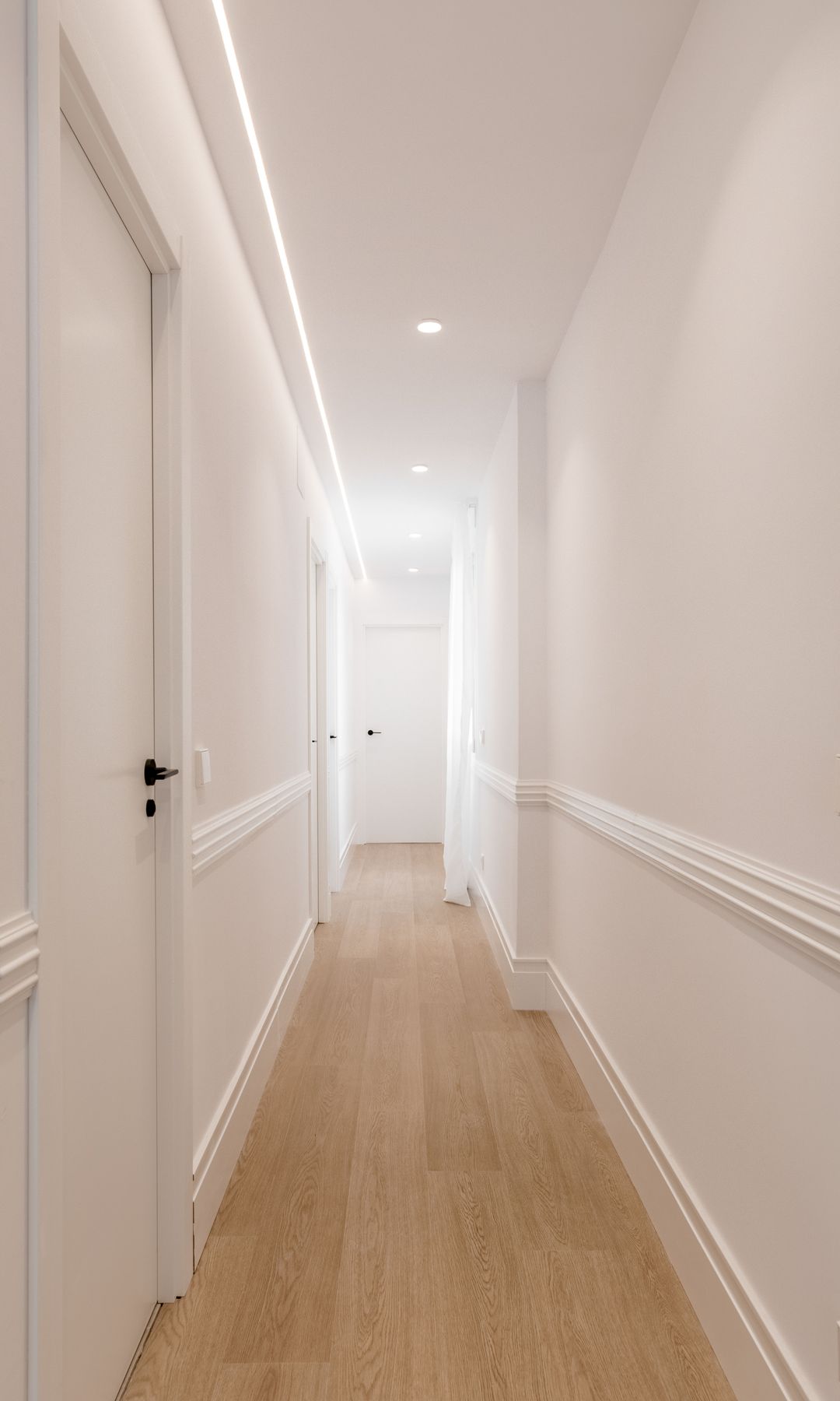 Pasillo blanco con zócalo alto con moldura, focos empotrado en el techo, suelo de madera claro 