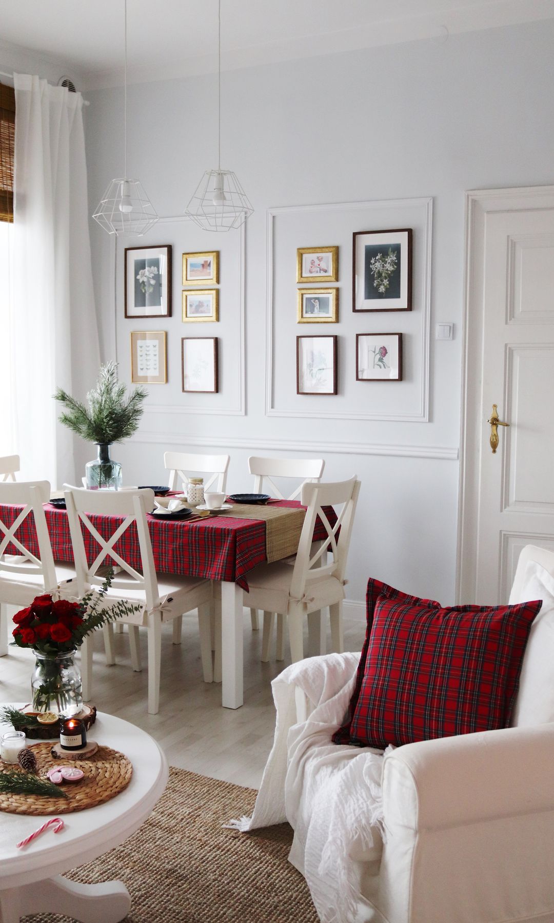 Comedor con molduras en las paredes, cuadros y fotos, mesa de comedor vestida de Navidad con mantel rojo, sillas blancas de madera, butaca con funda blanca, cojín rojo de cuadros, mesa de centro con velas y arreglo floral