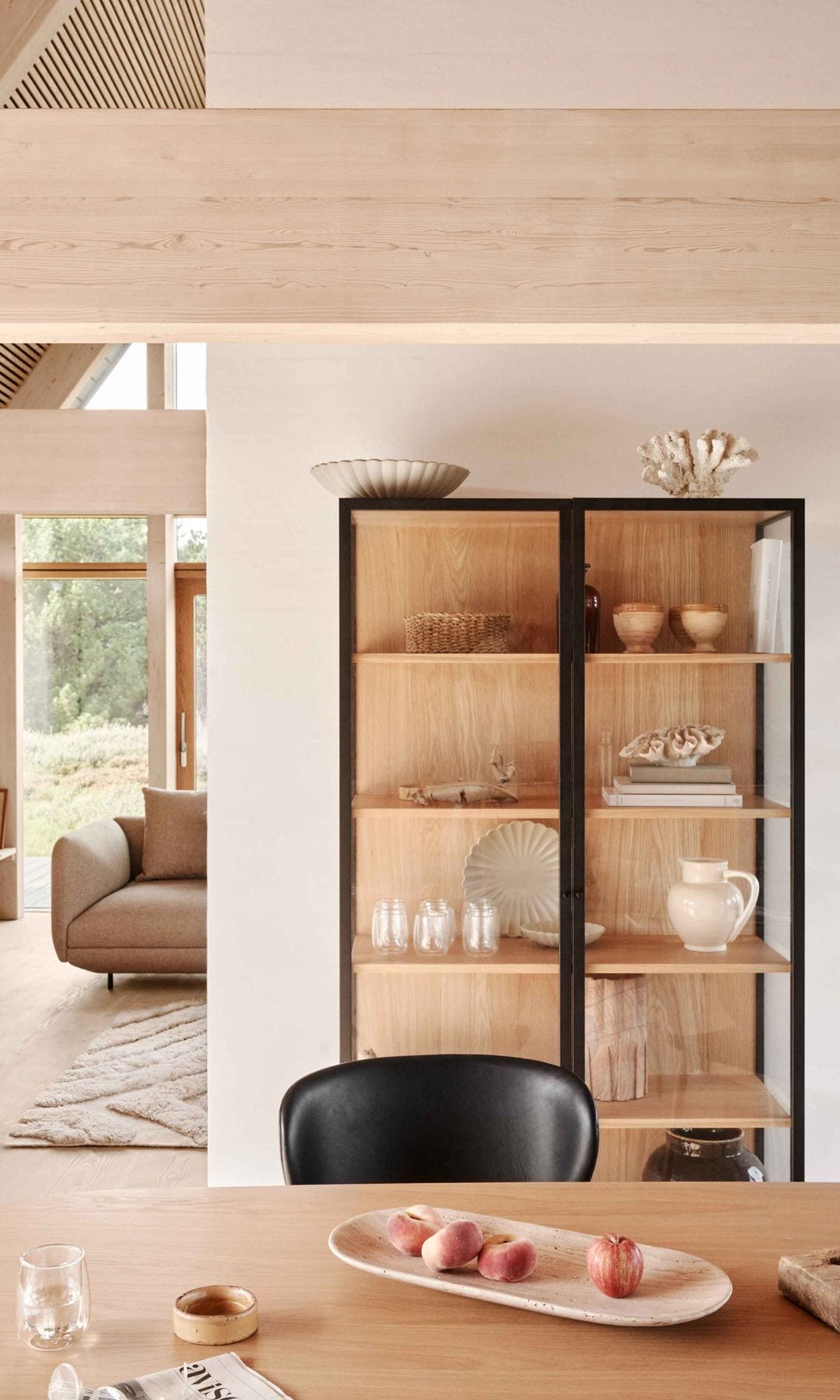 Salón comedor separados por medio tabique, vitrina con la trasera de madera y los perfiles negros, vajilla, copas, mesa de comedor de madera, sillas negras, sofá beige