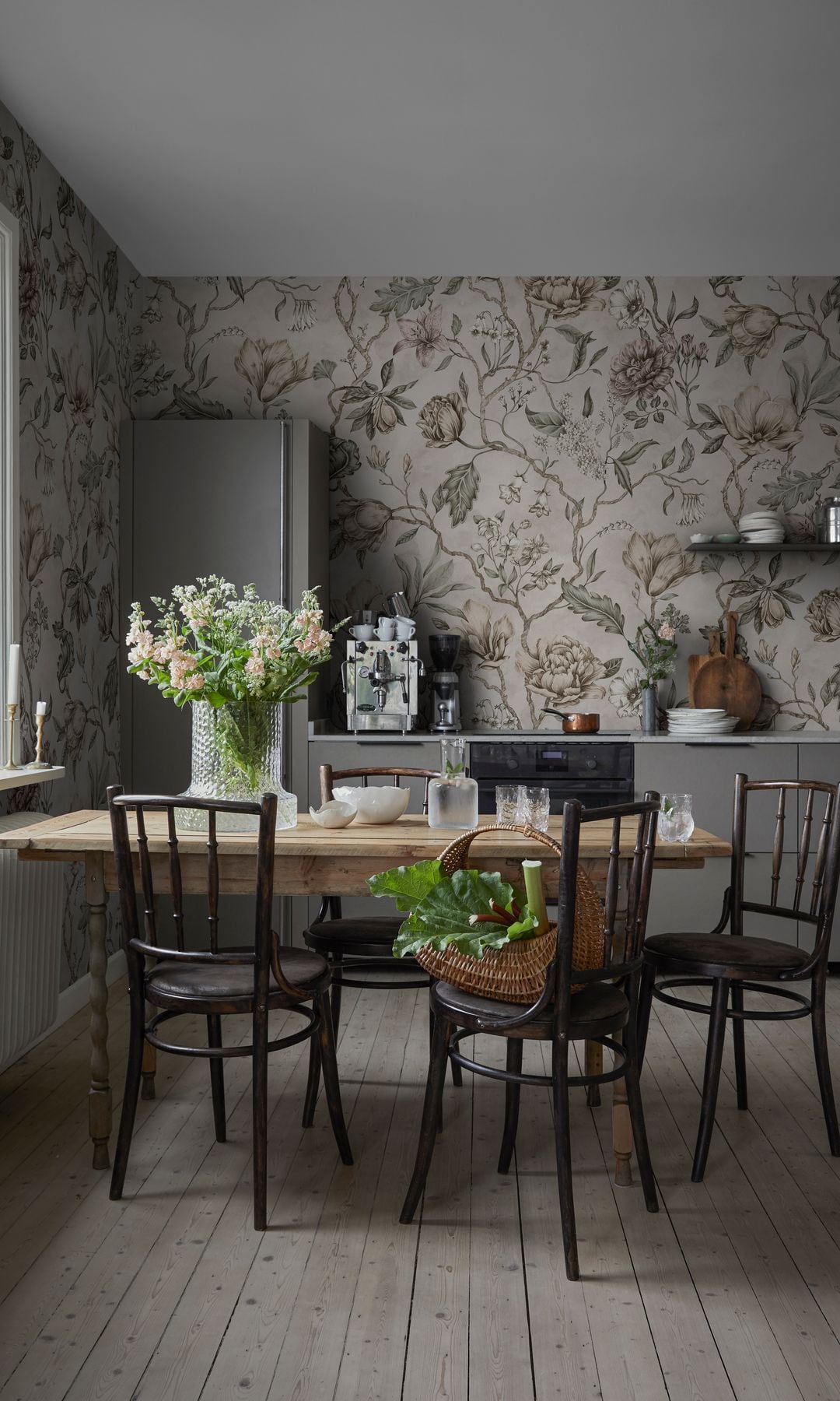 Cocina con papel pintado de motivos vegetales, mesa de madera, sillas de madera negra, jarrón con flores, cesto con verduras y suelo de madera