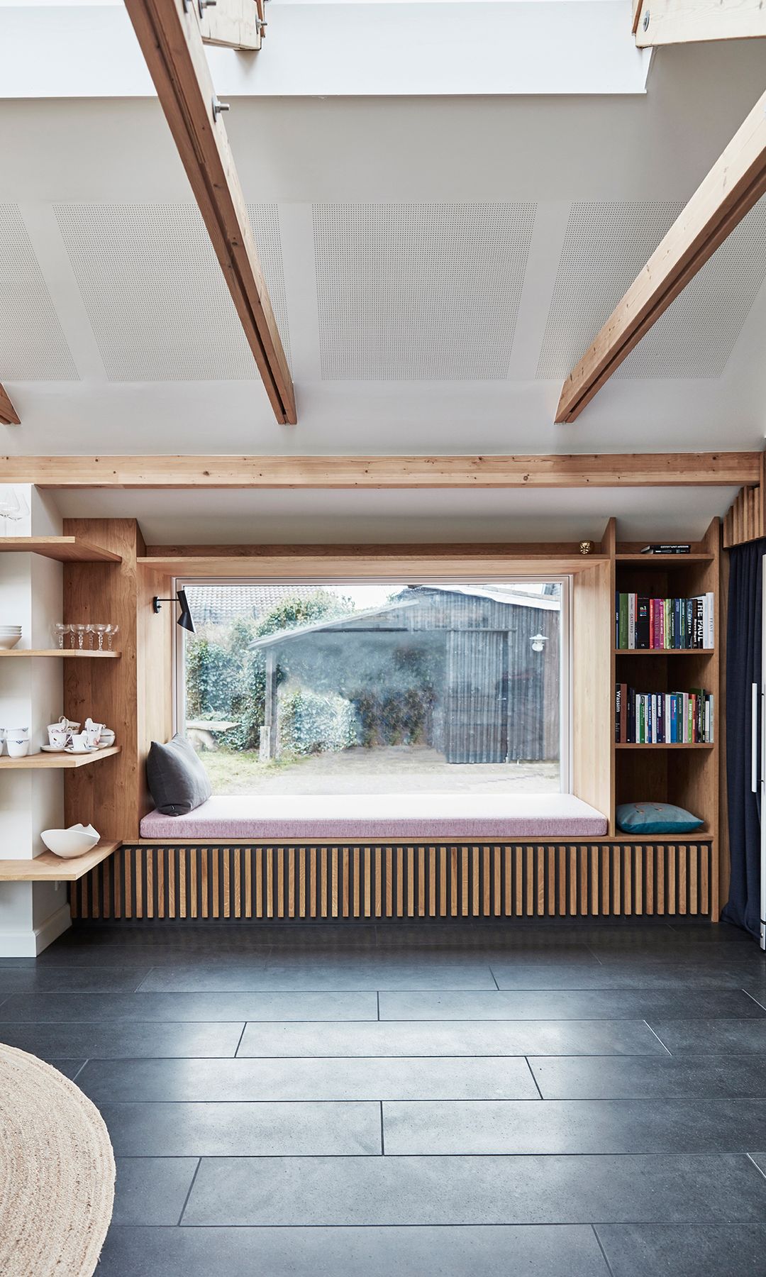 Rincón bajo la ventana con banco de listones de madera, colchoneta de color rosa, estantería a los lados, ventanal con vistas al jardín 