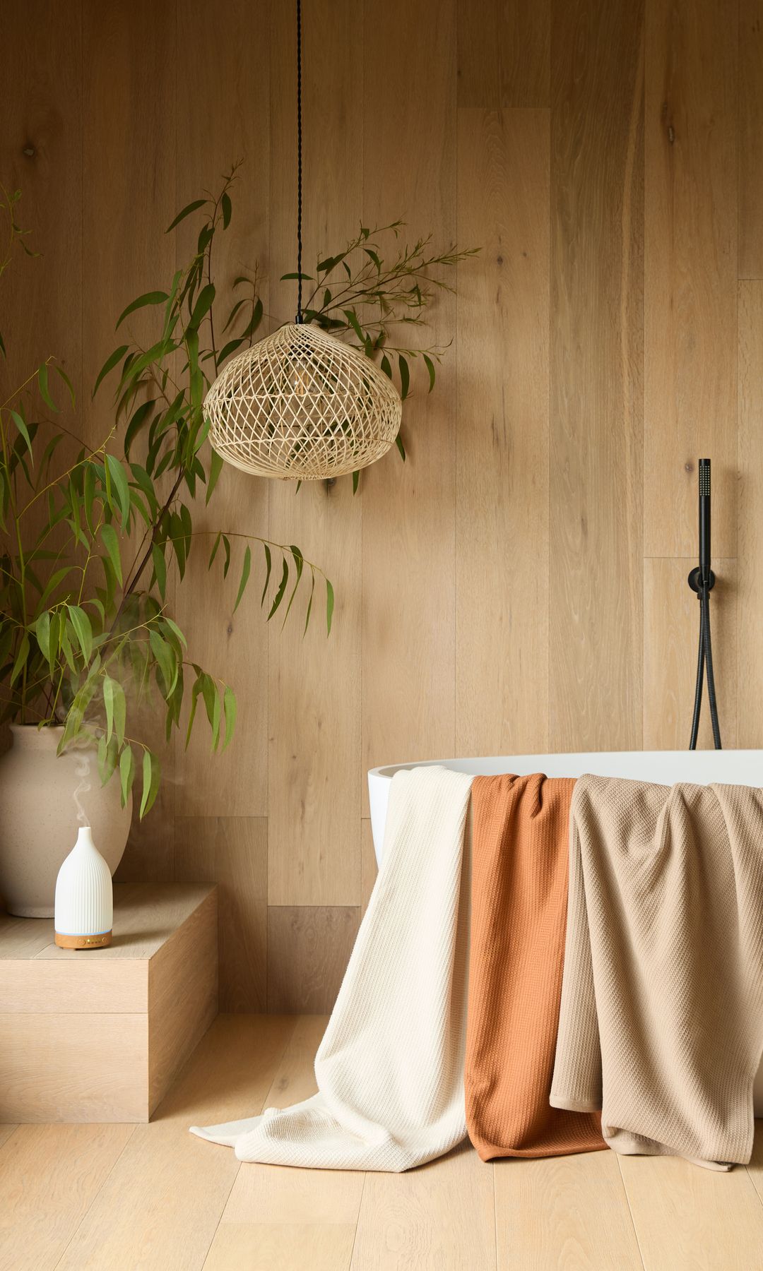 Baño con bañera exenta, toallas sobre la bañera, paredes y suelo de madera, lámpara de fibras vegetales, planta en maceta de barro 