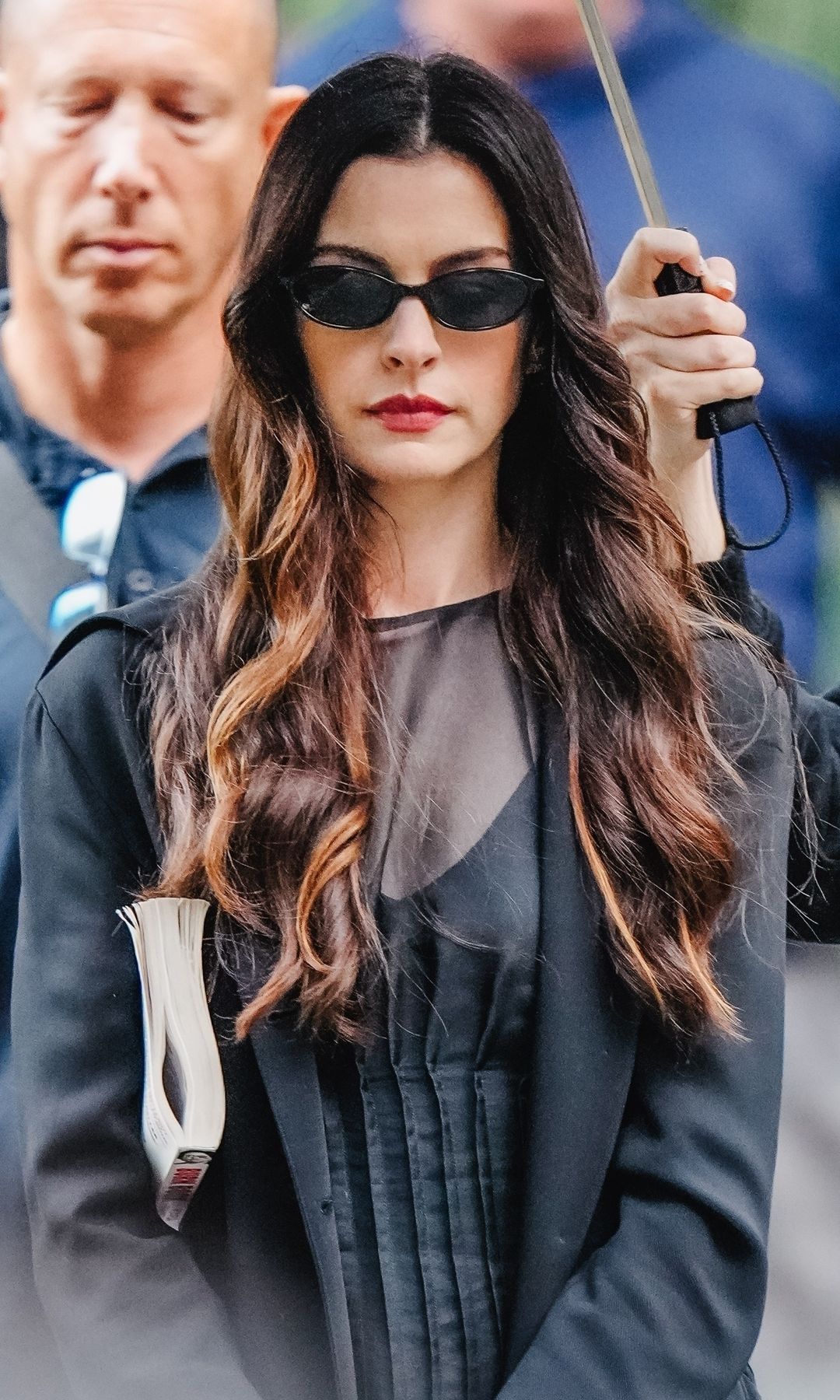 Anne Hathaway, con gafas de sol y abrigo negro, camina entre una multitud.