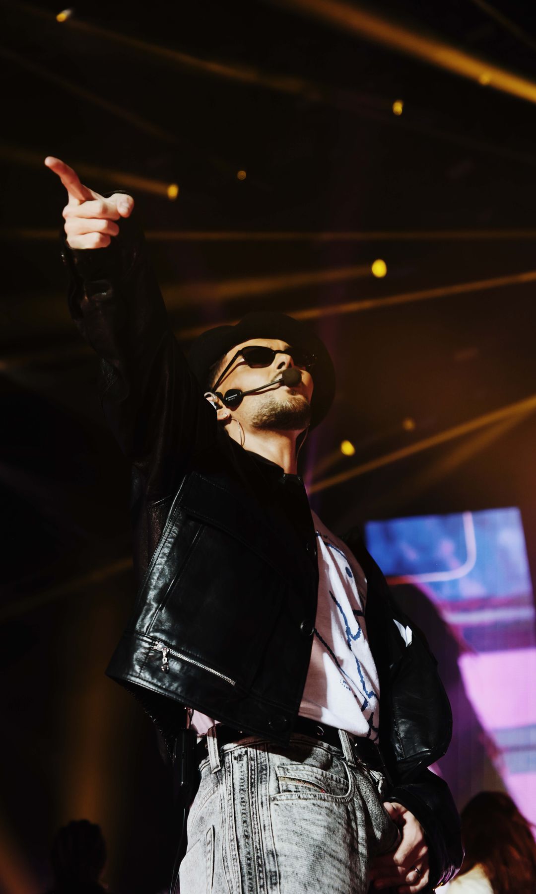 Abraham Mateo  en el escenario con chaqueta de cuero, gafas y micrófono, señalando.