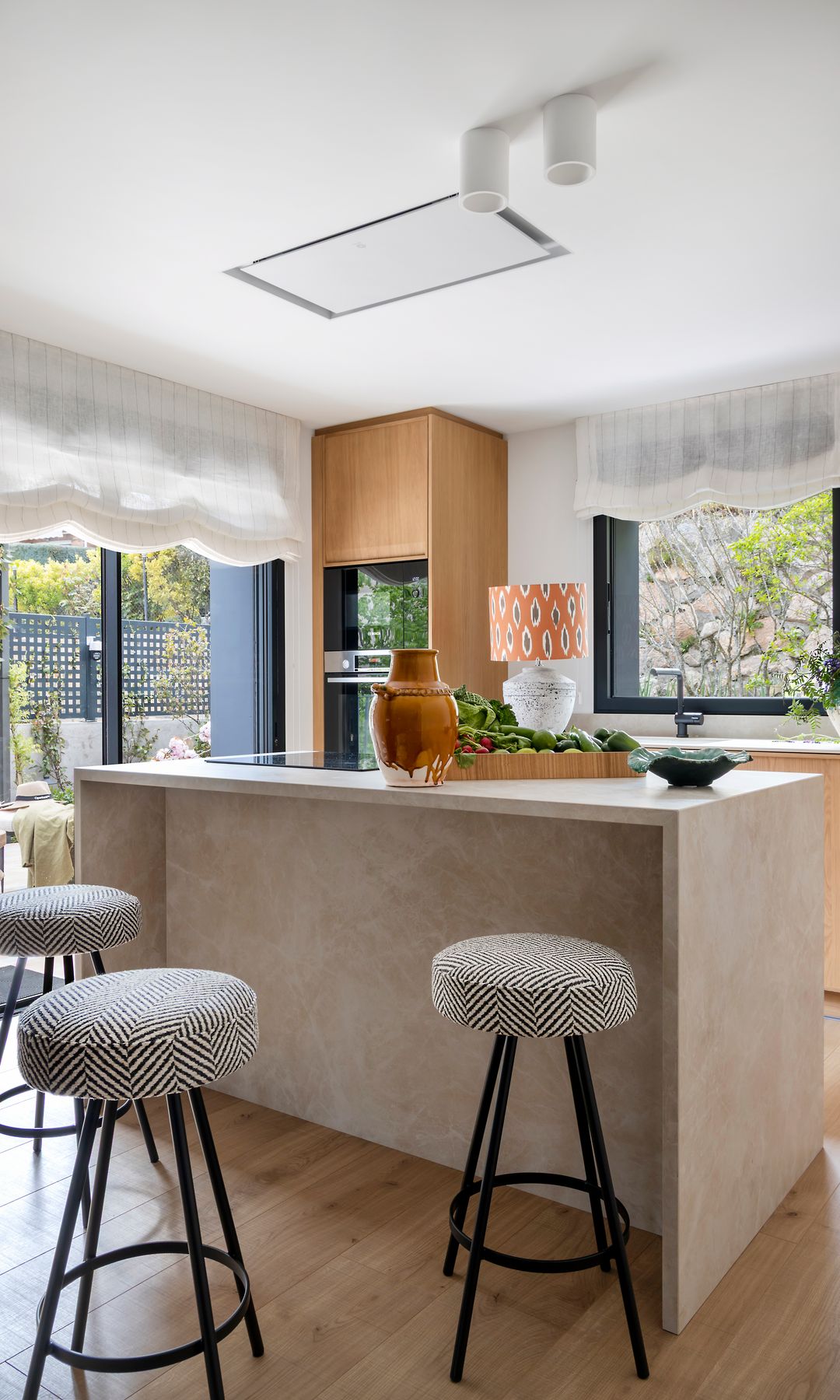 Cocina con isla con muebles de madera, taburetes altos, suelo de madera, salida a la terraza con estores 