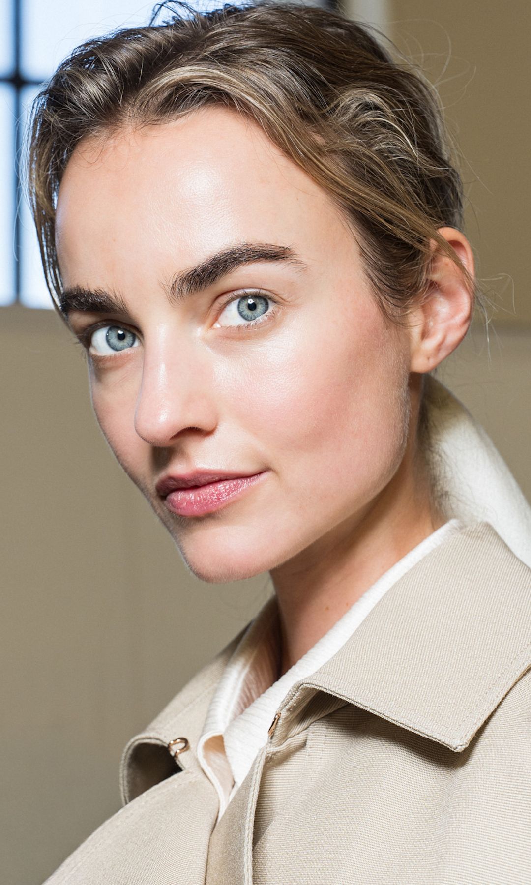 Modelo con ojos azules y recogido rizado, gabardina y camisa almidonada en el backstage de Gabriela Hearst