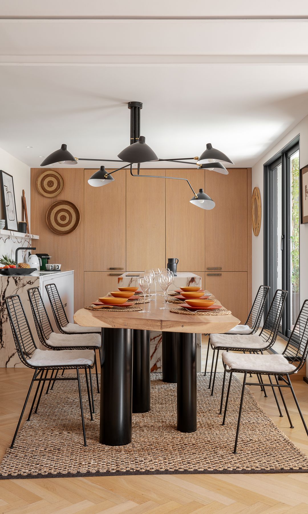 Comedor con mesa de madera con patas negras, alfombra de fibras vegetales, armario de madera XL, lámpara de techo de color negro, sillas de comedor metálicas negras, cuadros en la pared