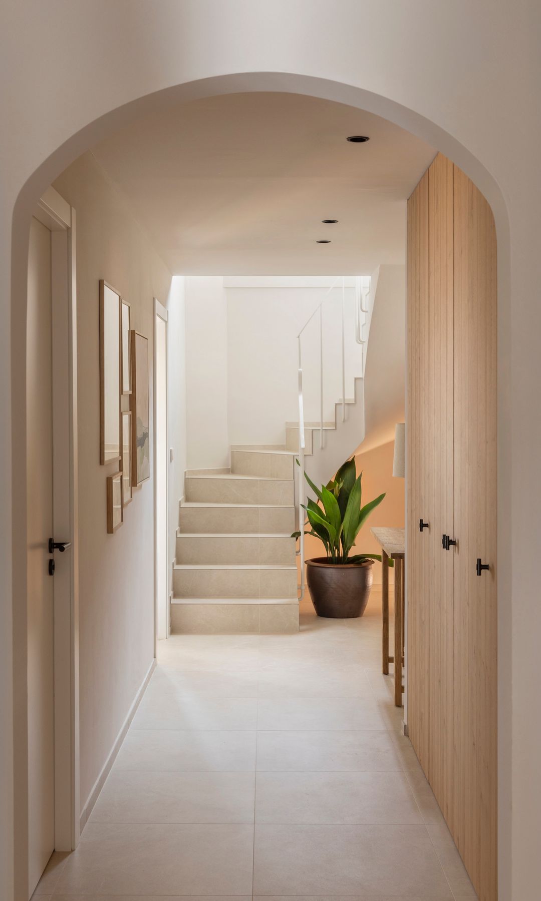 Pasillo con pared de armarios de madera clara, arco al final y escaleras blancas en la otra parte, maceta con planta 